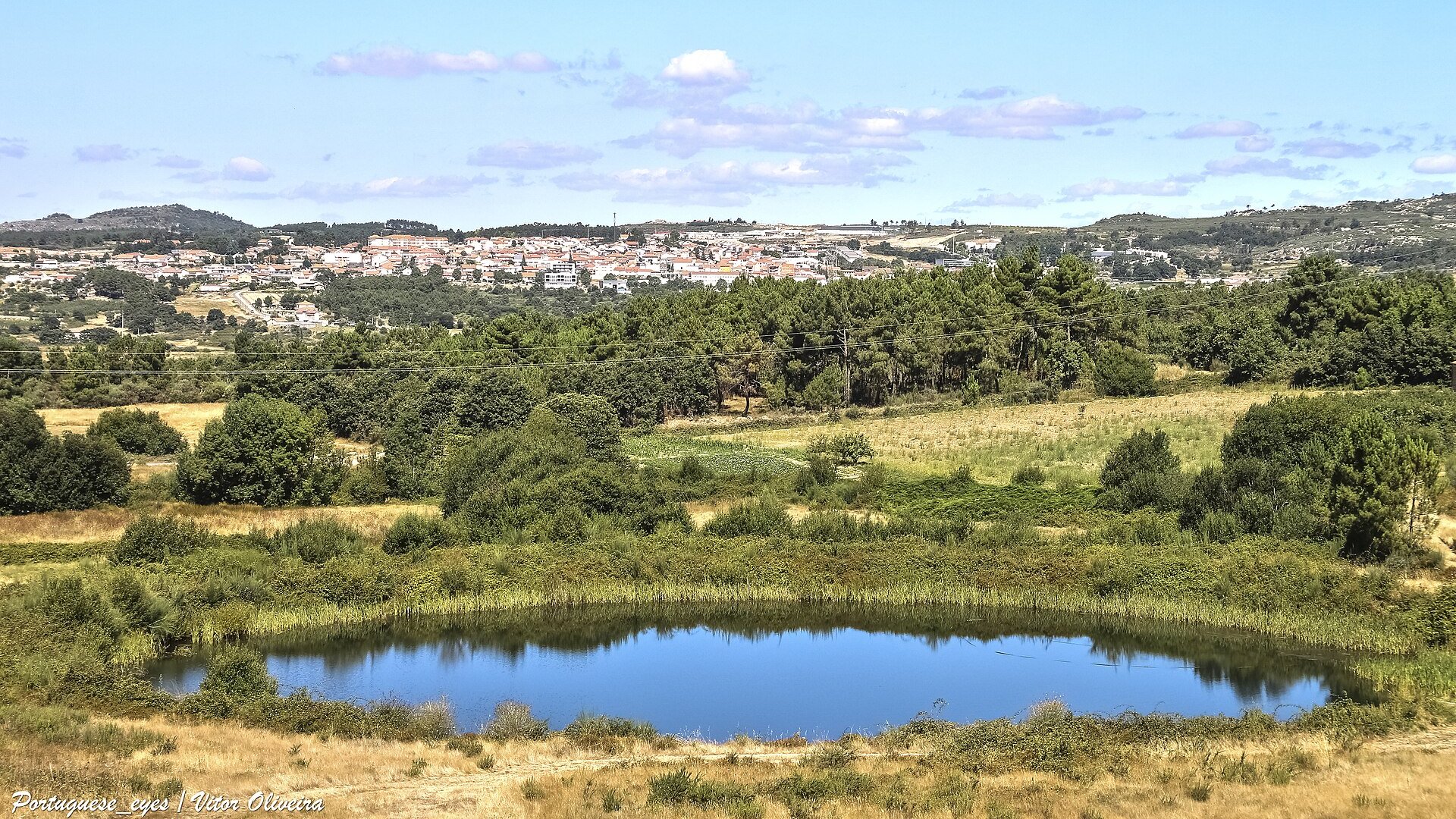 Vista da vila de Carrazeda de Ansiães, no alto de uma encosta, a partir de um miradouro: pequeno açude em primeiro plano, pinhal e campos secos a meio, casario branco de telha vermelha na linha do horizonte