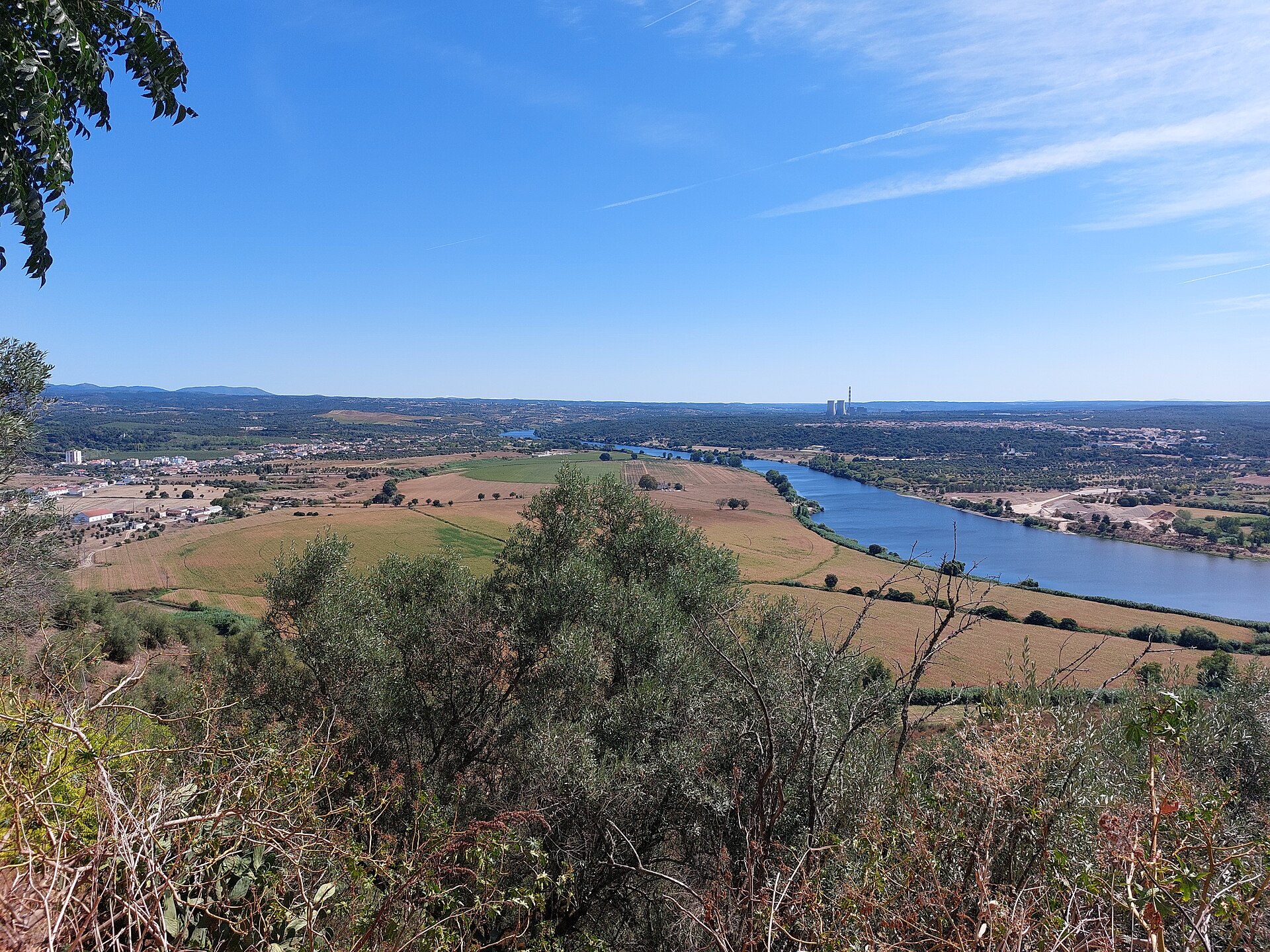 Vista elevada sobre o vale do Tejo, com campos agrícolas, o rio ao centro e, ao fundo, as torres e a chaminé da Central Termoeléctrica do Pego entre colinas arborizadas