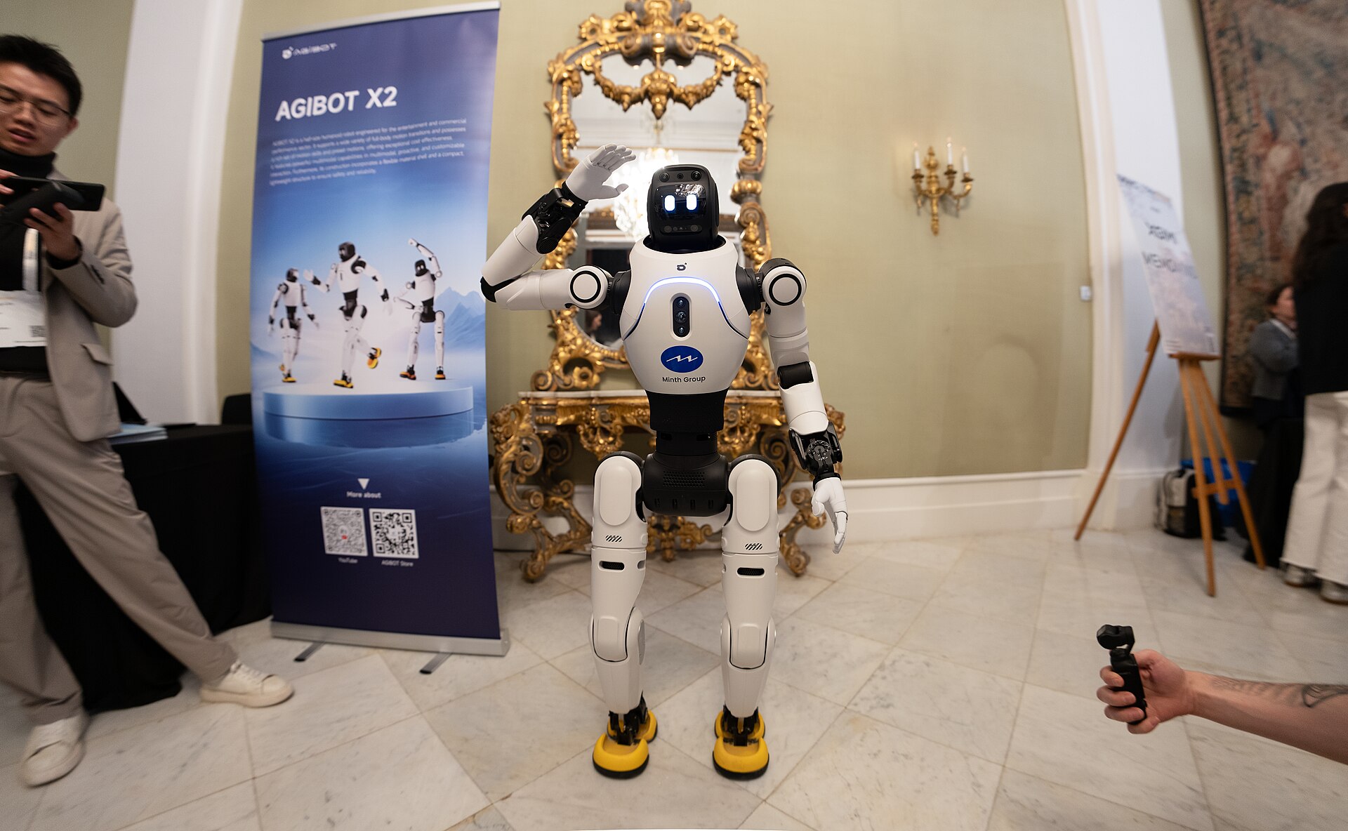 Robô humanoide branco e preto com o logótipo Minth Group no peito e um braço levantado, em frente a uma bandeira azul «AGIBOT X2», num salão com espelho dourado.