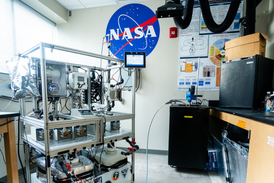 Rig experimental de biorreator de circuito fechado com tubos, sensores e ecrã de controlo, em laboratório com logo da NASA