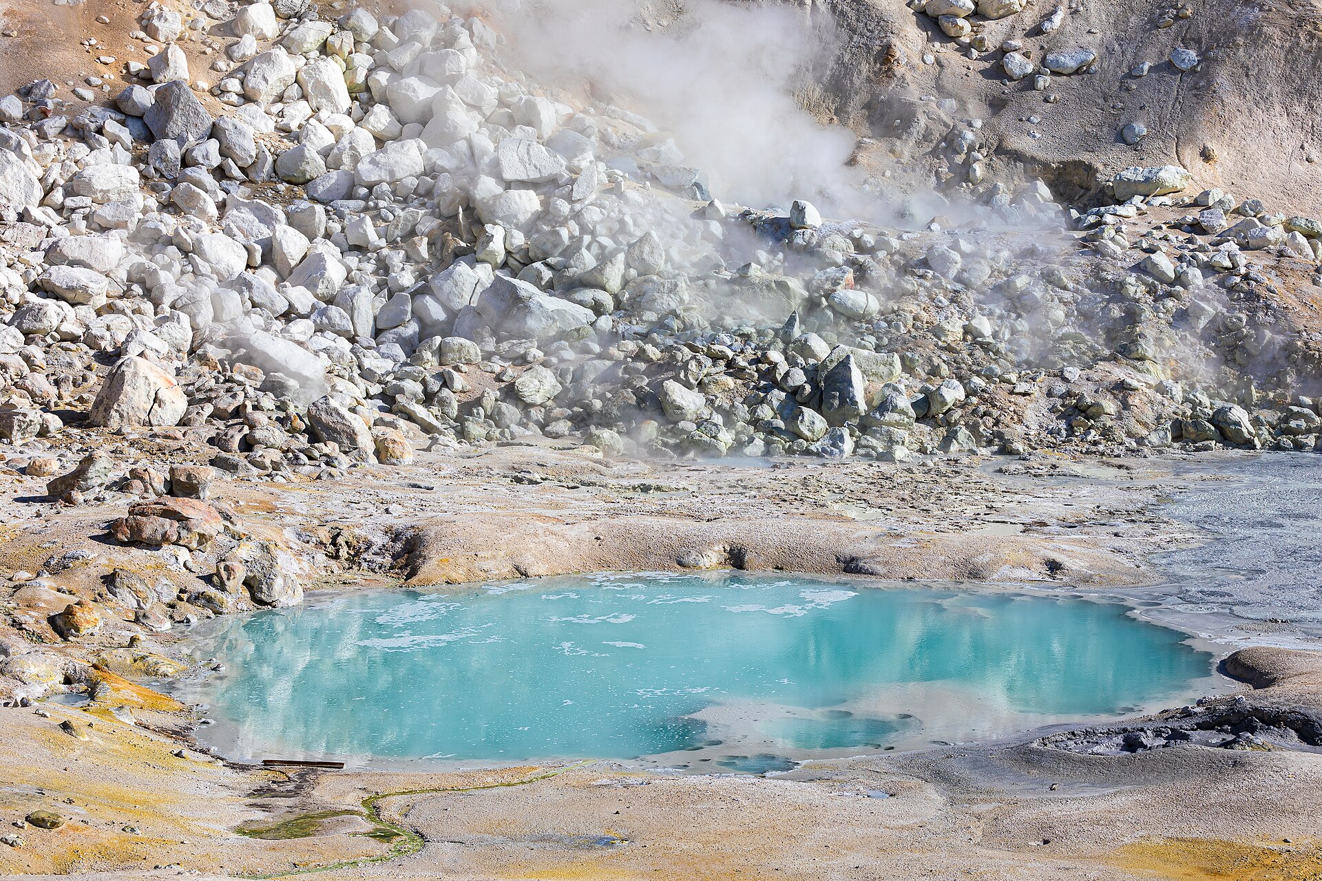 Poça de água azul-turquesa rodeada de rochas brancas e acinzentadas, com vapor a sair do solo em volta