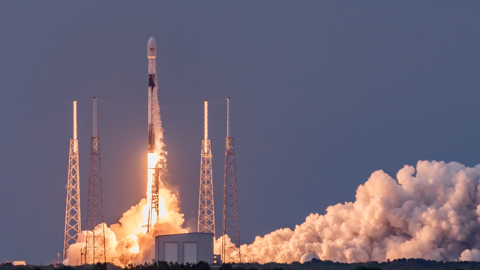 Falcon 9 a descolar do SLC-40, no Cabo Canaveral, ao amanhecer: o foguetão branco sobe entre quatro torres metálicas, com uma enorme nuvem de vapor iluminada de laranja pelo escape, sobre um céu azul-escuro.