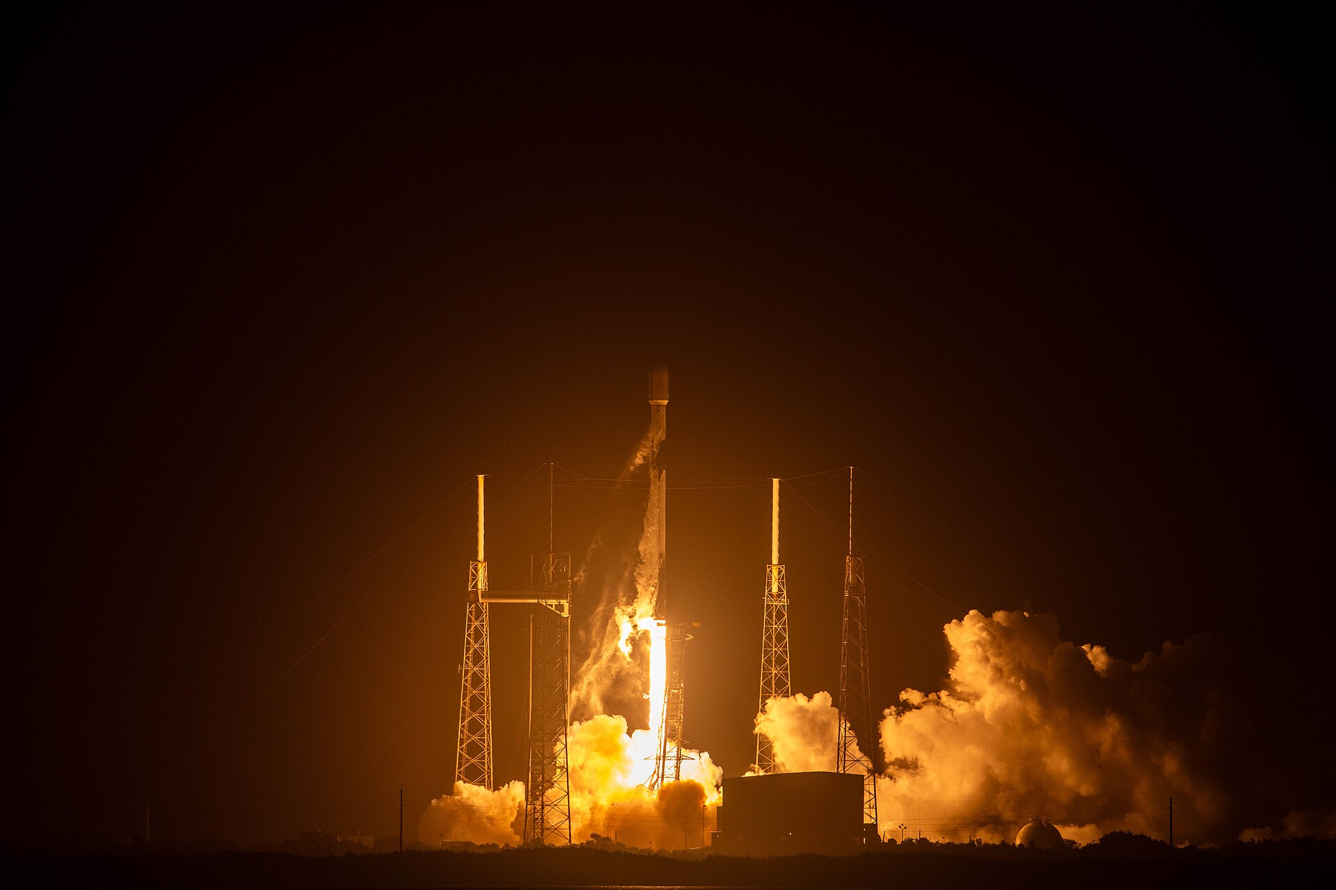 Falcon 9 a descolar de noite de Cape Canaveral com chama intensa e torres de serviço