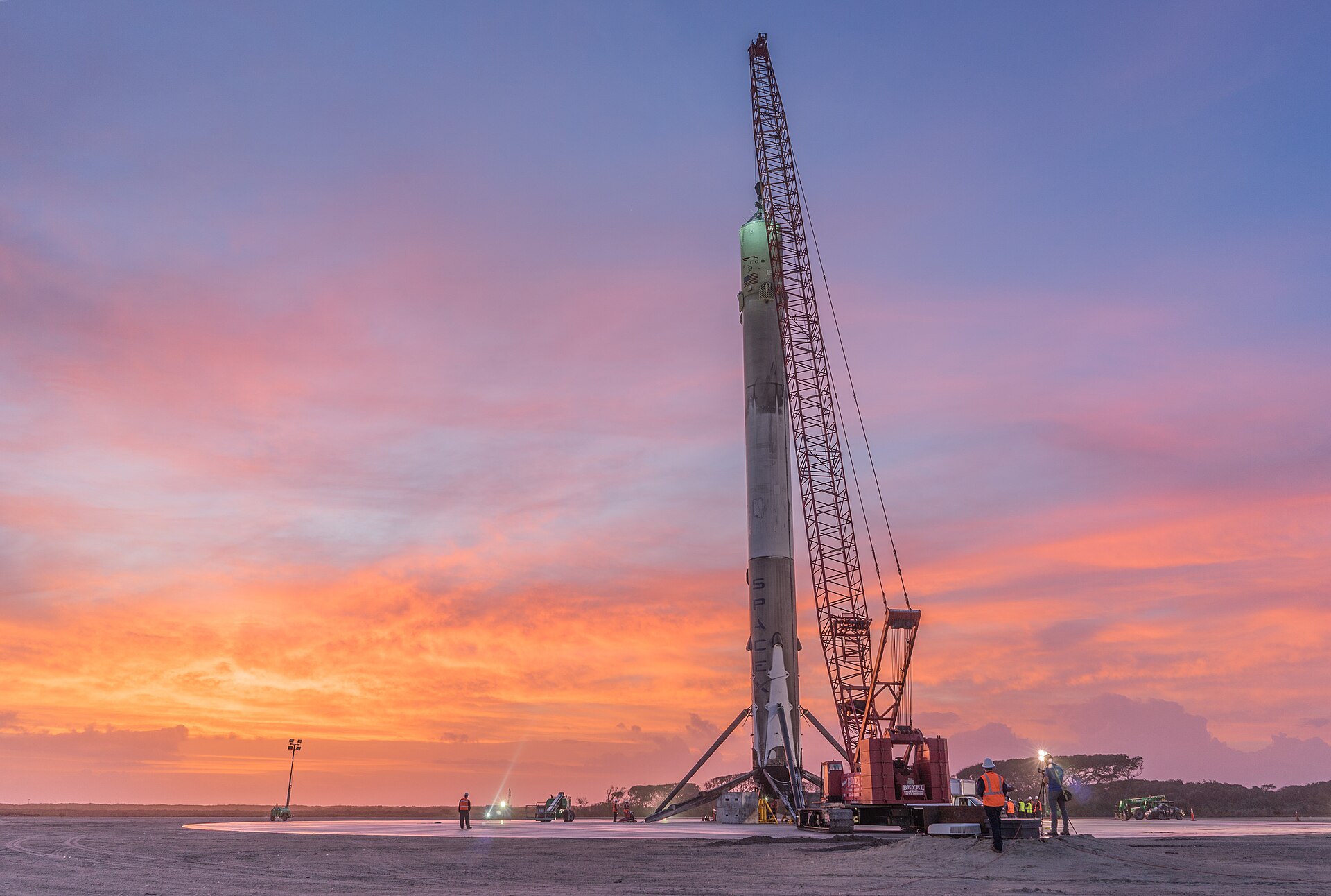 Primeiro estágio Falcon 9 da SpaceX no solo após aterragem, ao pôr do sol, com grua e técnicos