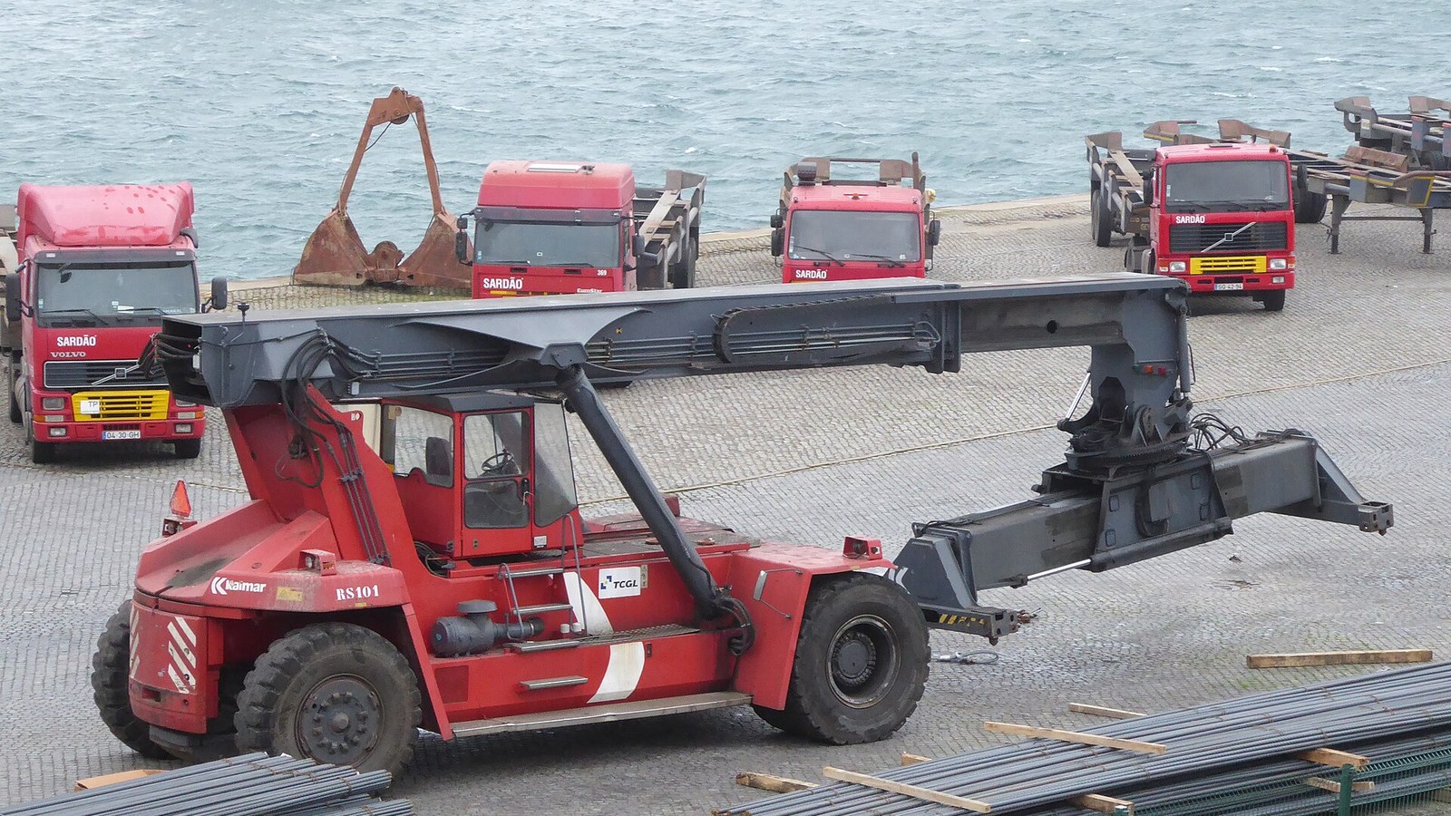Um reach stacker vermelho da marca Kalmar, com a lança levantada e o spreader suspenso, num cais de pavimento de calçada junto à água; atrás, quatro camiões vermelhos com a inscrição SARDÃO e, em primeiro plano, varões de aço empilhados.