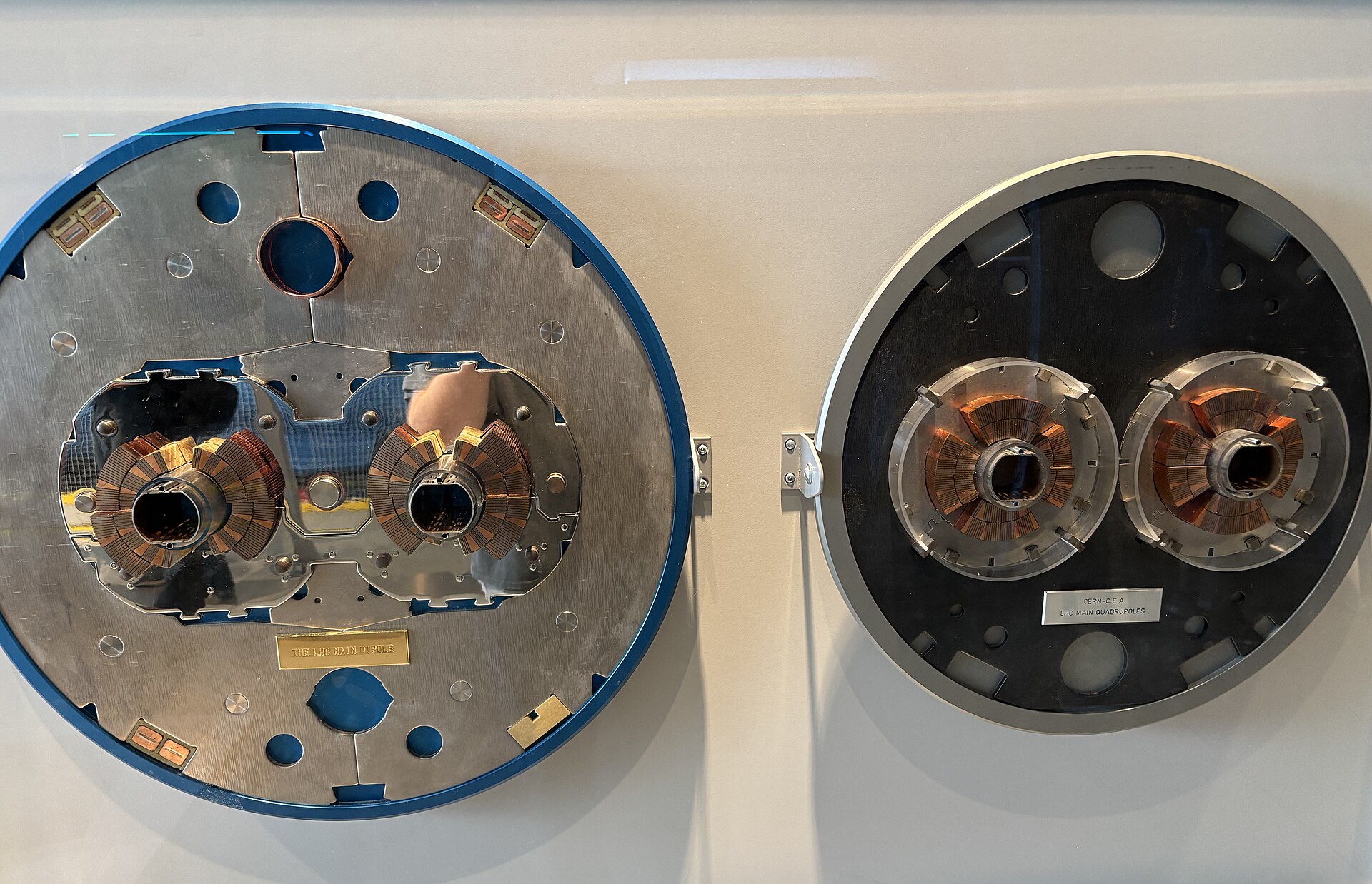 Duas secções circulares de ímanes do LHC expostas numa parede: à esquerda, o dipolo principal, com dois enrolamentos de cobre em torno de dois tubos; à direita, o quadrupolo principal, com dois enrolamentos de cobre em forma de anel