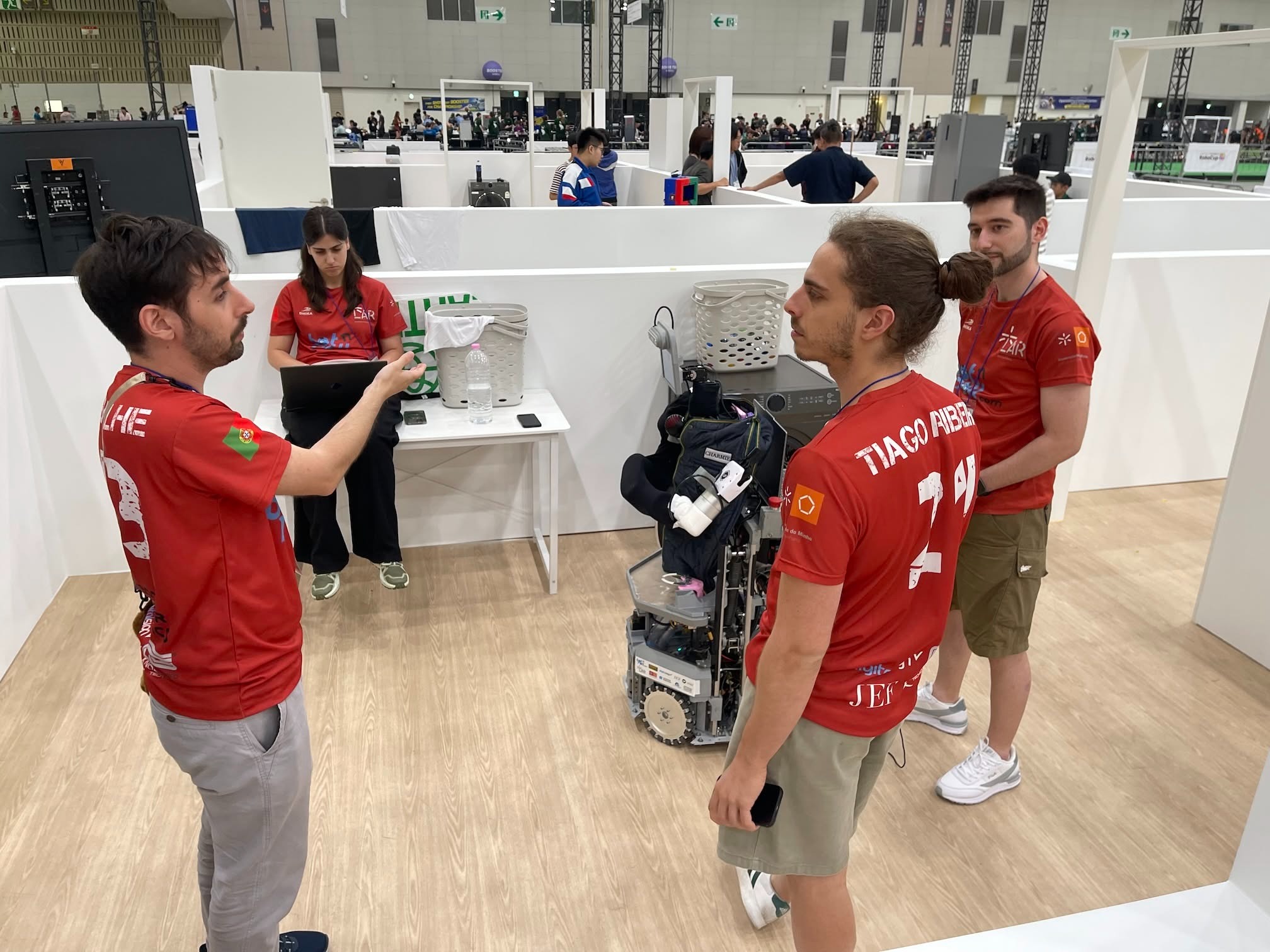 O robô CHARMIE da Universidade do Minho em ação durante as provas da RoboCup@Home 2026, em Incheon, Coreia do Sul. O braço manipulador branco é visível, com a equipa a monitorizar o desempenho ao fundo.