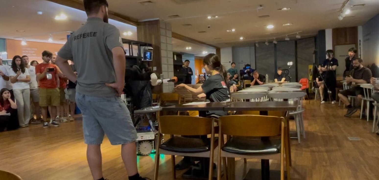 O robô CHARMIE numa prova de interação com um avaliador durante a RoboCup@Home 2026. A equipa e o juiz observam o desempenho do robô numa simulação de ambiente doméstico.