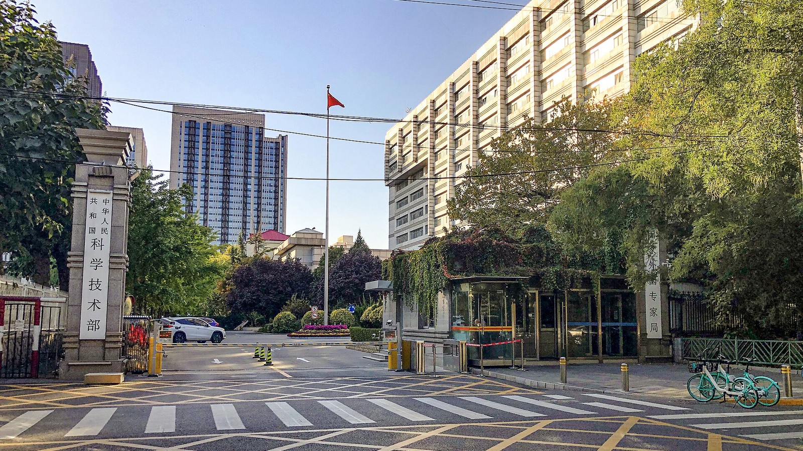 Entrada de um ministério em Pequim: pilares de pedra com inscrições verticais em caracteres chineses à esquerda e à direita, portaria envidraçada, mastro com bandeira vermelha ao centro, edifícios altos ao fundo, passadeira e marcas rodoviárias amarelas no pavimento e bicicletas partilhadas estacionadas.