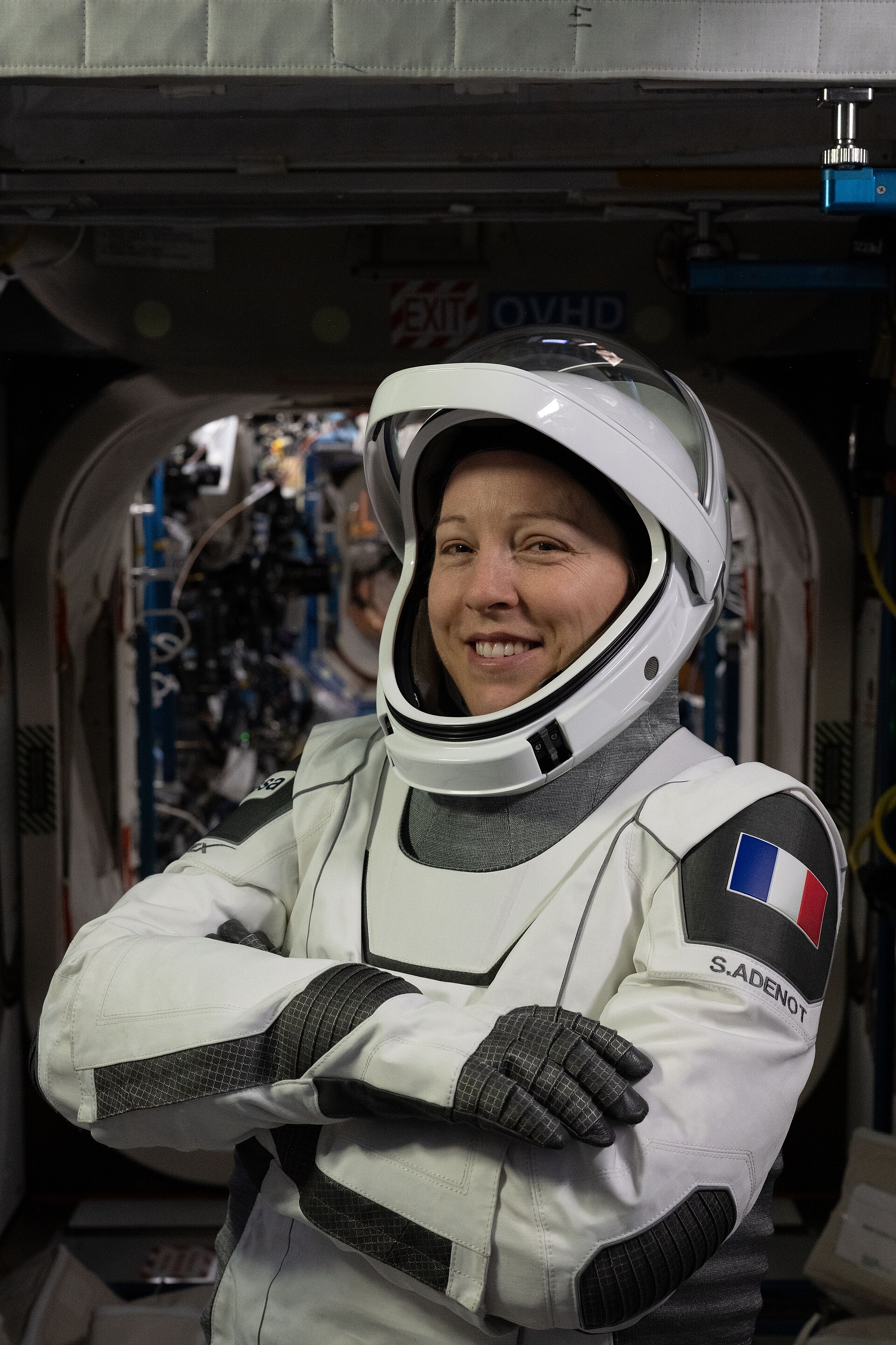 A astronauta Sophie Adenot, com o fato de pressão branco da SpaceX e o capacete aberto, de braços cruzados no interior da Estação Espacial Internacional; no braço esquerdo vêem-se a bandeira francesa e a etiqueta S.ADENOT.