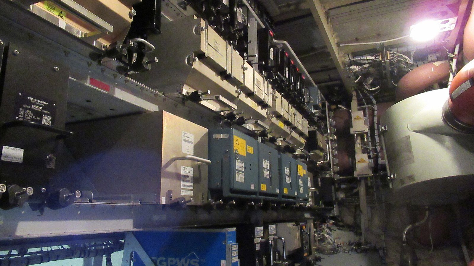 Sala de aviónica de um Airbus A319 vista de perto: várias filas de computadores de bordo cinzentos e pretos encaixados em suportes com pegas e rótulos, cablagem em feixes, uma caixa azul identificada com a sigla GPWS e equipamento rotulado vibro-meter