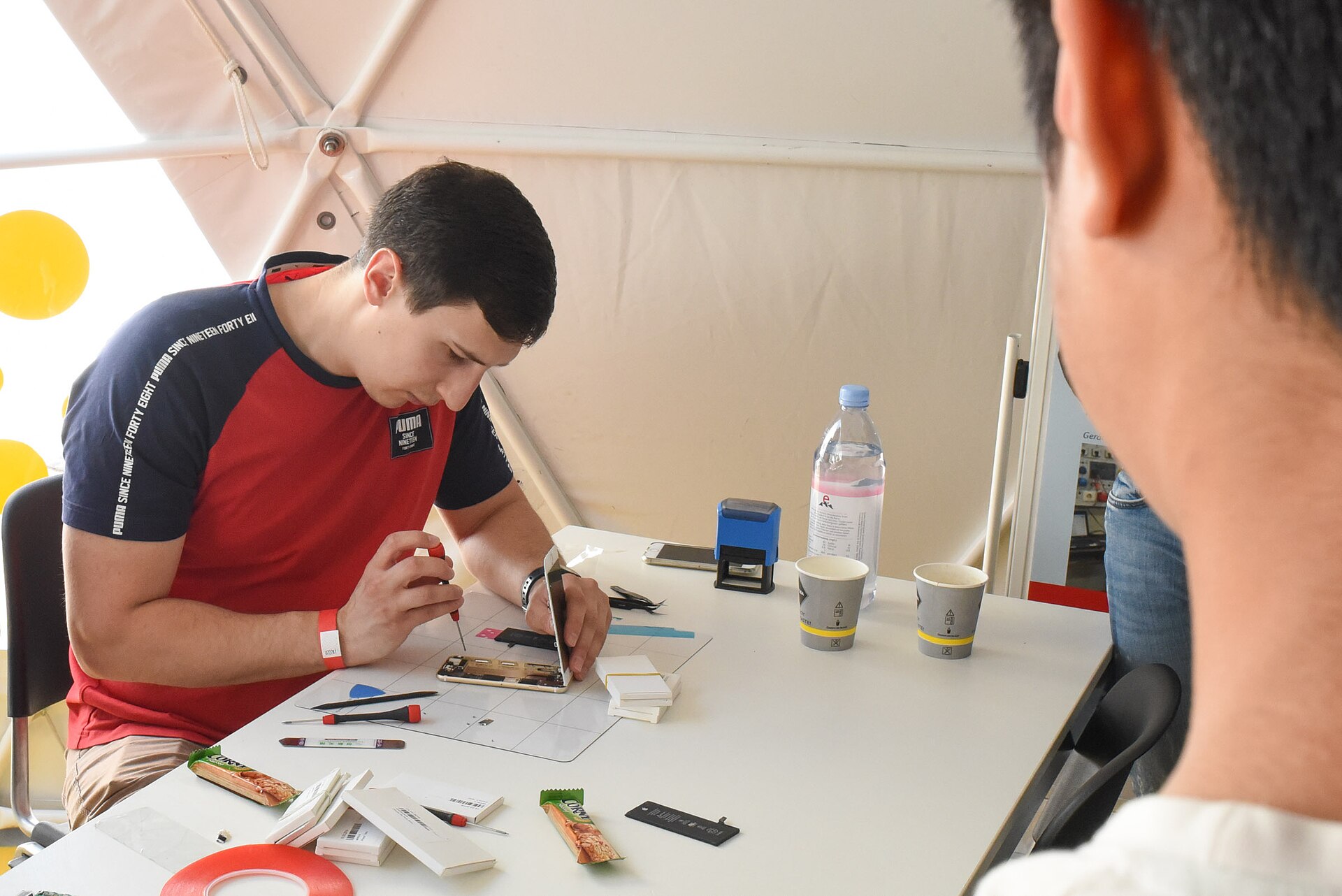 Jovem de camisola vermelha e azul, sentado a uma mesa branca, abre um telemóvel com uma chave de precisão; sobre a mesa estão espátulas, lâminas, cartões e fita adesiva