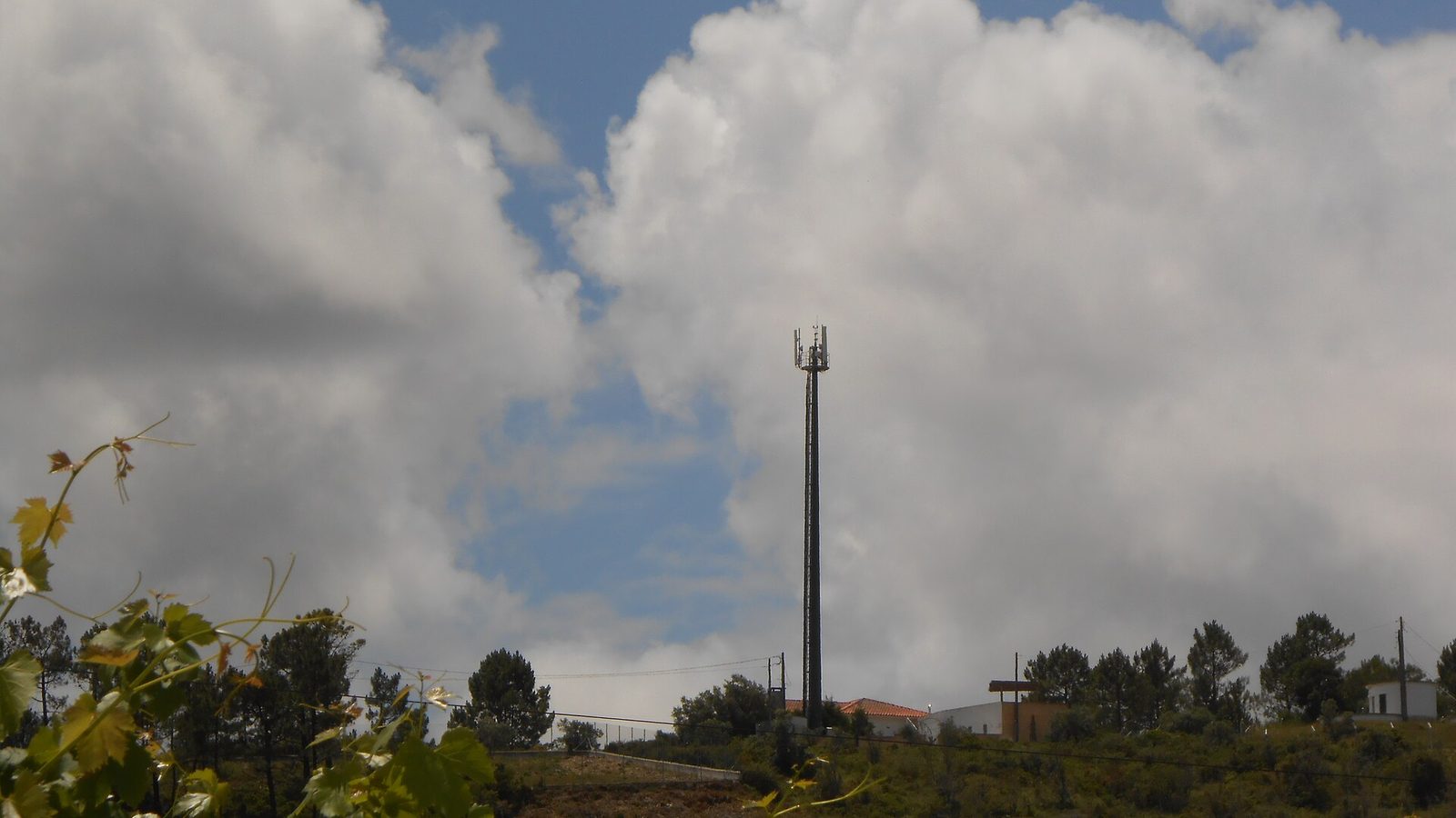 Antena de rede móvel: mastro escuro com painéis de antenas no topo, junto a um recinto vedado com edifícios baixos, sobre uma encosta com árvores e céu com nuvens.