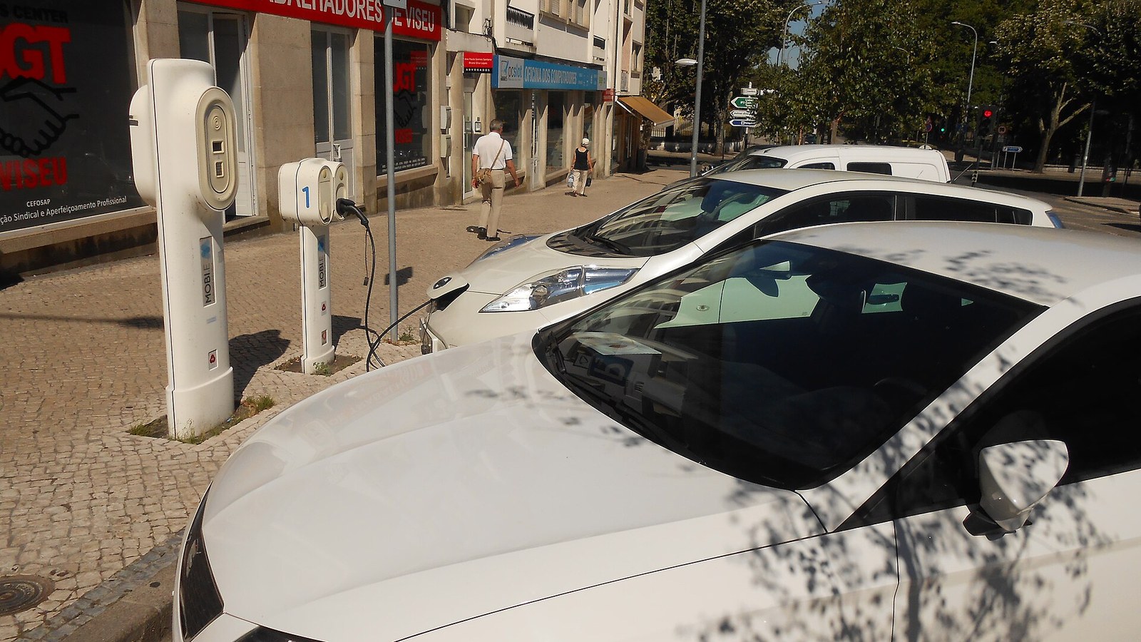 Dois automóveis elétricos brancos ligados por cabo a postos de carregamento Mobi.E numa rua de Viseu, com peões no passeio e edifícios ao fundo
