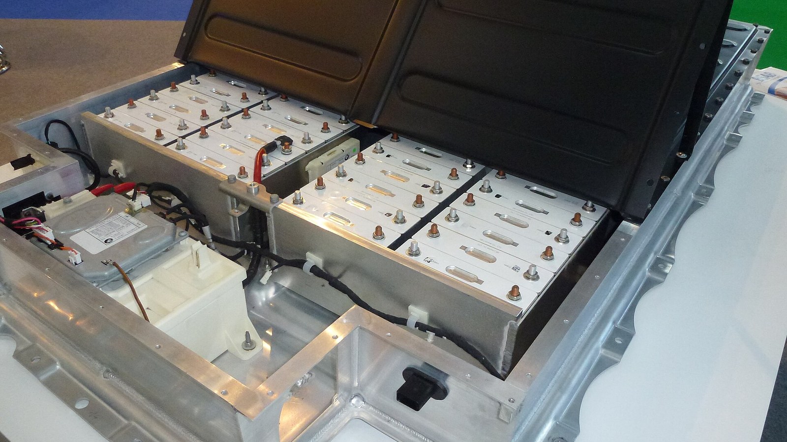 Bateria de tração de um automóvel elétrico aberta numa bancada, com módulos retangulares de células, terminais e cabos laranja e pretos, dentro de uma bandeja de alumínio