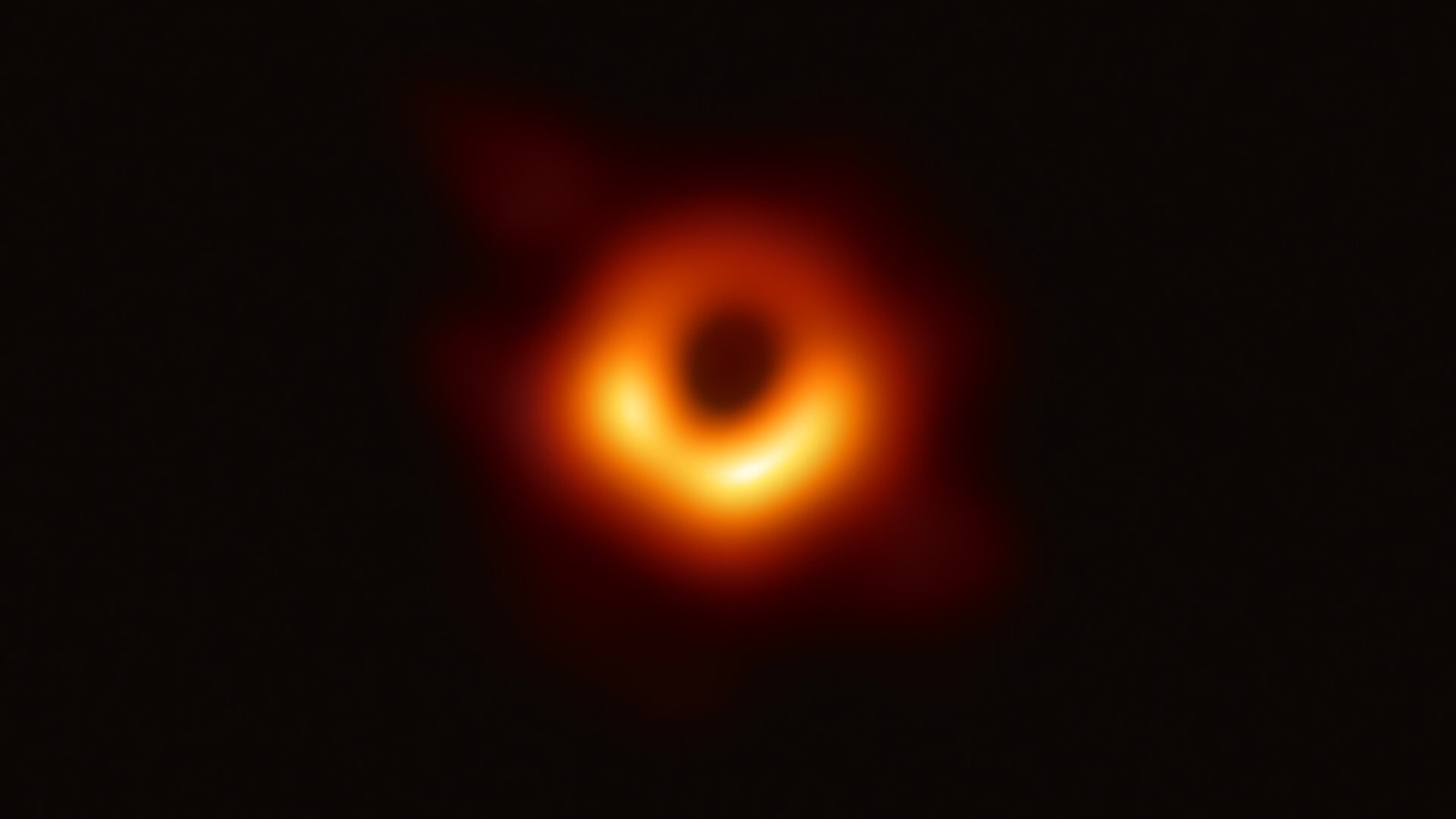 Imagem do buraco negro M87*: uma sombra escura circular rodeada por um anel laranja e amarelo irregular, sobre fundo negro