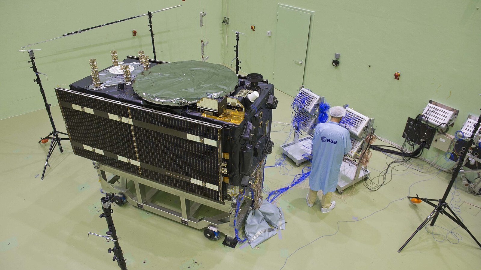 Satélite Galileo em configuração de voo numa sala de ensaios de paredes verdes, com painéis e coberturas de protecção, e um técnico de bata azul identificada com a sigla ESA a trabalhar junto de equipamento de teste e cabos