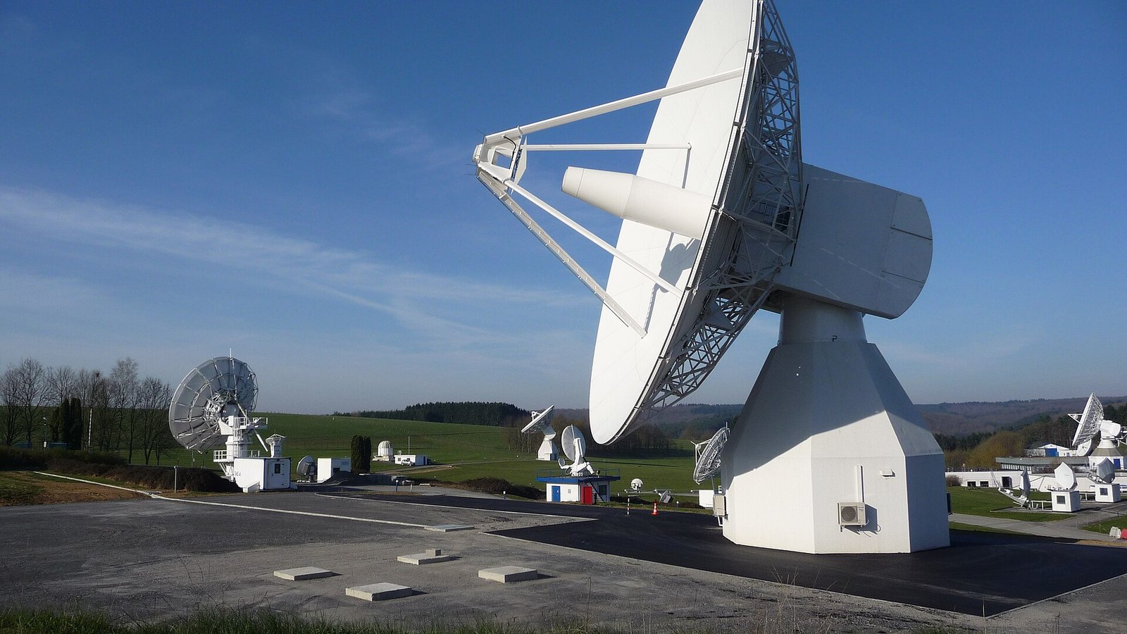 Grande antena parabólica branca de banda L montada sobre uma base de betão, com outras antenas e um edifício técnico em segundo plano, num planalto verde sob céu azul