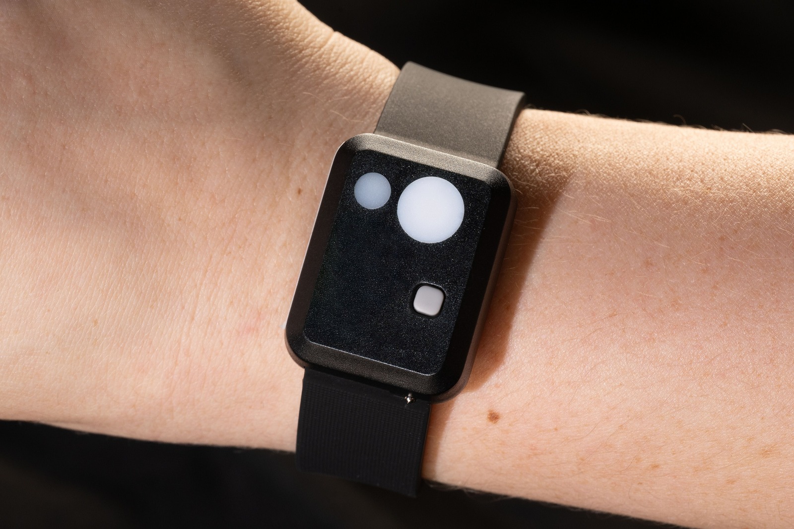 Primeiro plano de um pulso humano com um actígrafo de pulso preto, um pequeno aparelho rectangular com sensor redondo, preso por uma bracelete