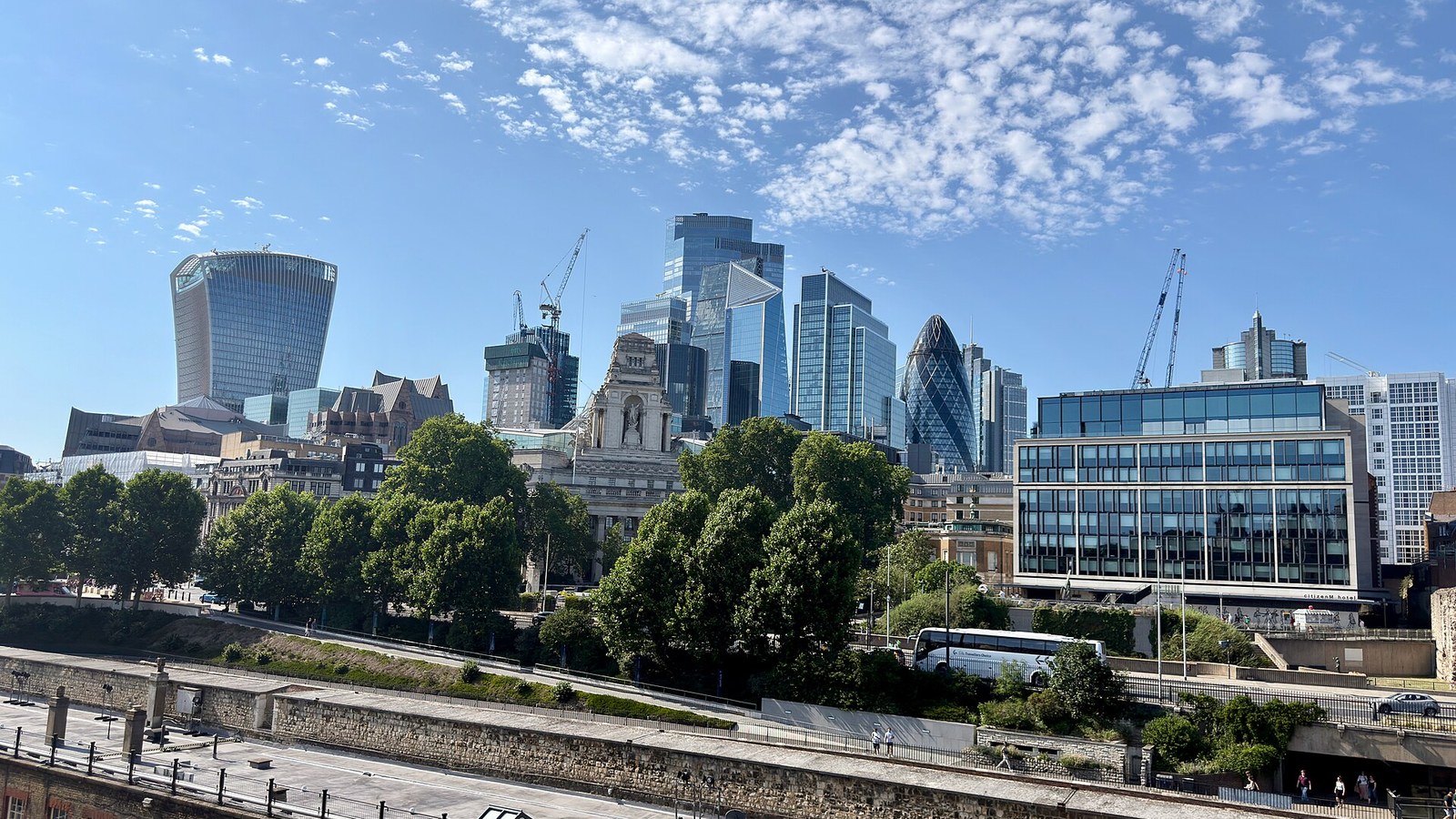 Panorama da City de Londres visto das muralhas da Torre de Londres, com os arranha-céus do distrito financeiro, gruas de obra e as muralhas de pedra em primeiro plano, sob um céu azul.