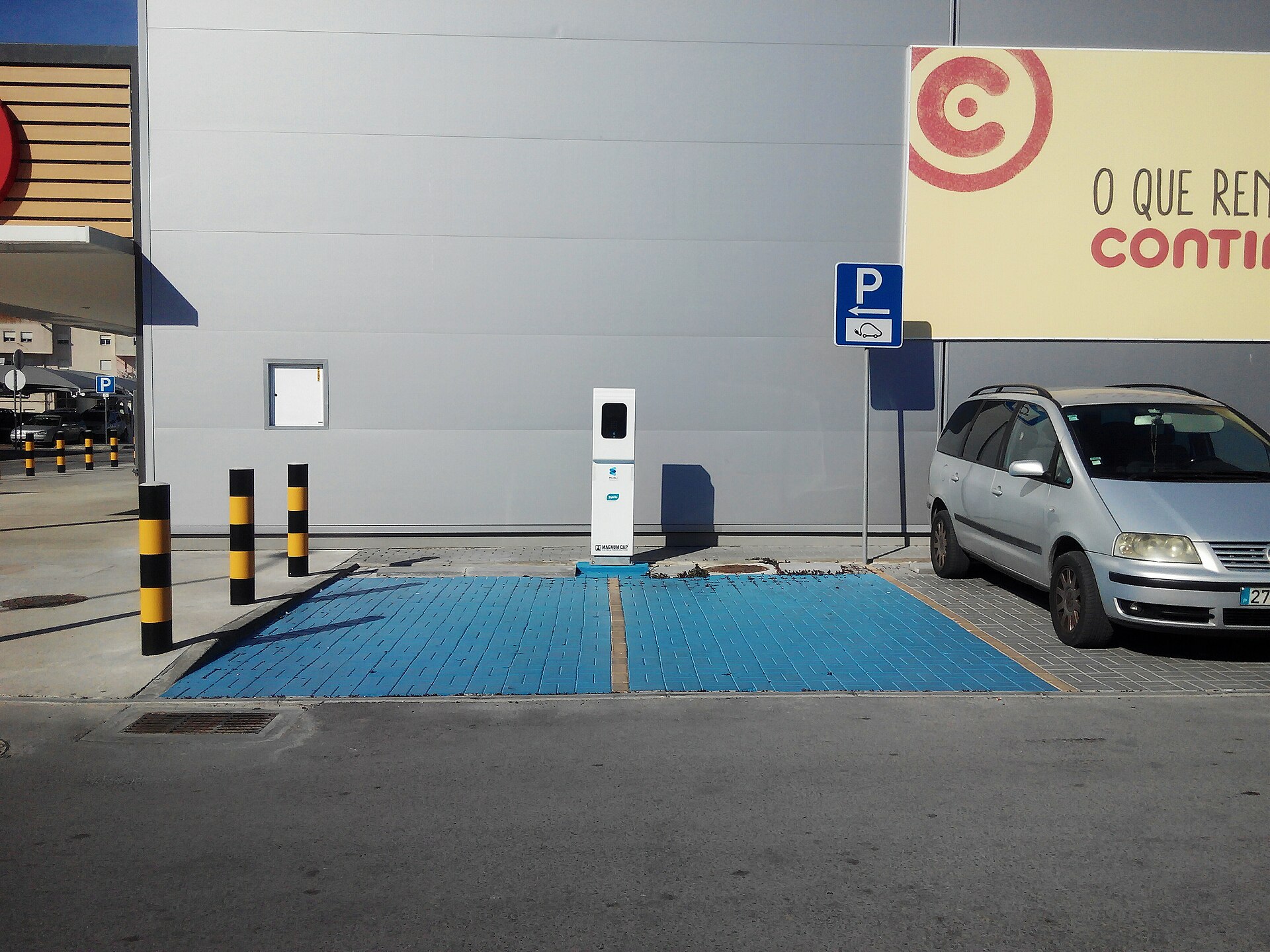 Posto de carregamento elétrico branco entre duas praças de estacionamento pintadas de azul, num parque de supermercado; à esquerda, pilaretes pretos e amarelos e um painel «P» com símbolo de veículo elétrico; à direita, uma carrinha cinzenta estacionada num lugar comum.