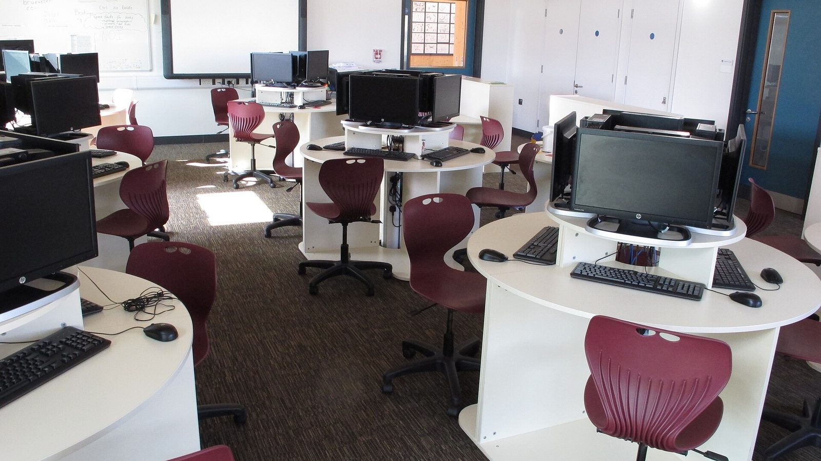 Sala de computadores de uma escola: secretárias brancas redondas com monitores, teclados e ratos, cadeiras escolares vermelhas e um quadro branco ao fundo.