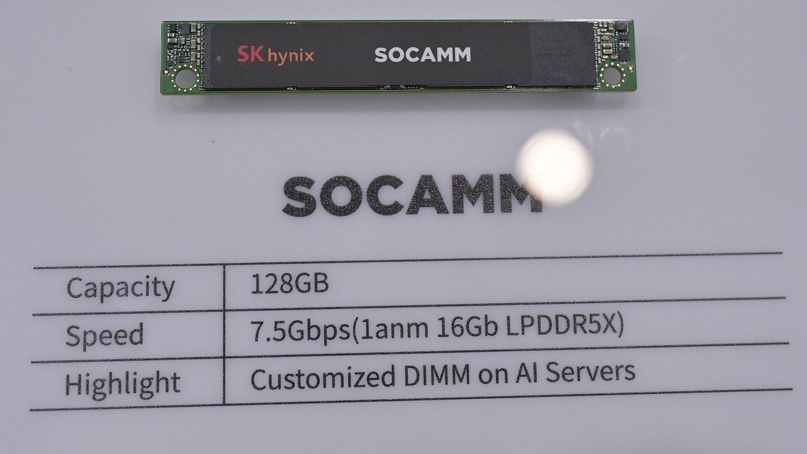 Módulo de memória alongado sobre placa verde, com o logótipo SK hynix e a palavra SOCAMM na etiqueta preta, sobre uma superfície branca; abaixo, impressas no mostrador, uma tabela com capacidade de 128 GB, velocidade de 7,5 Gbps em LPDDR5X e a indicação «Customized DIMM on AI Servers», e a palavra SOCAMM em letras grandes.