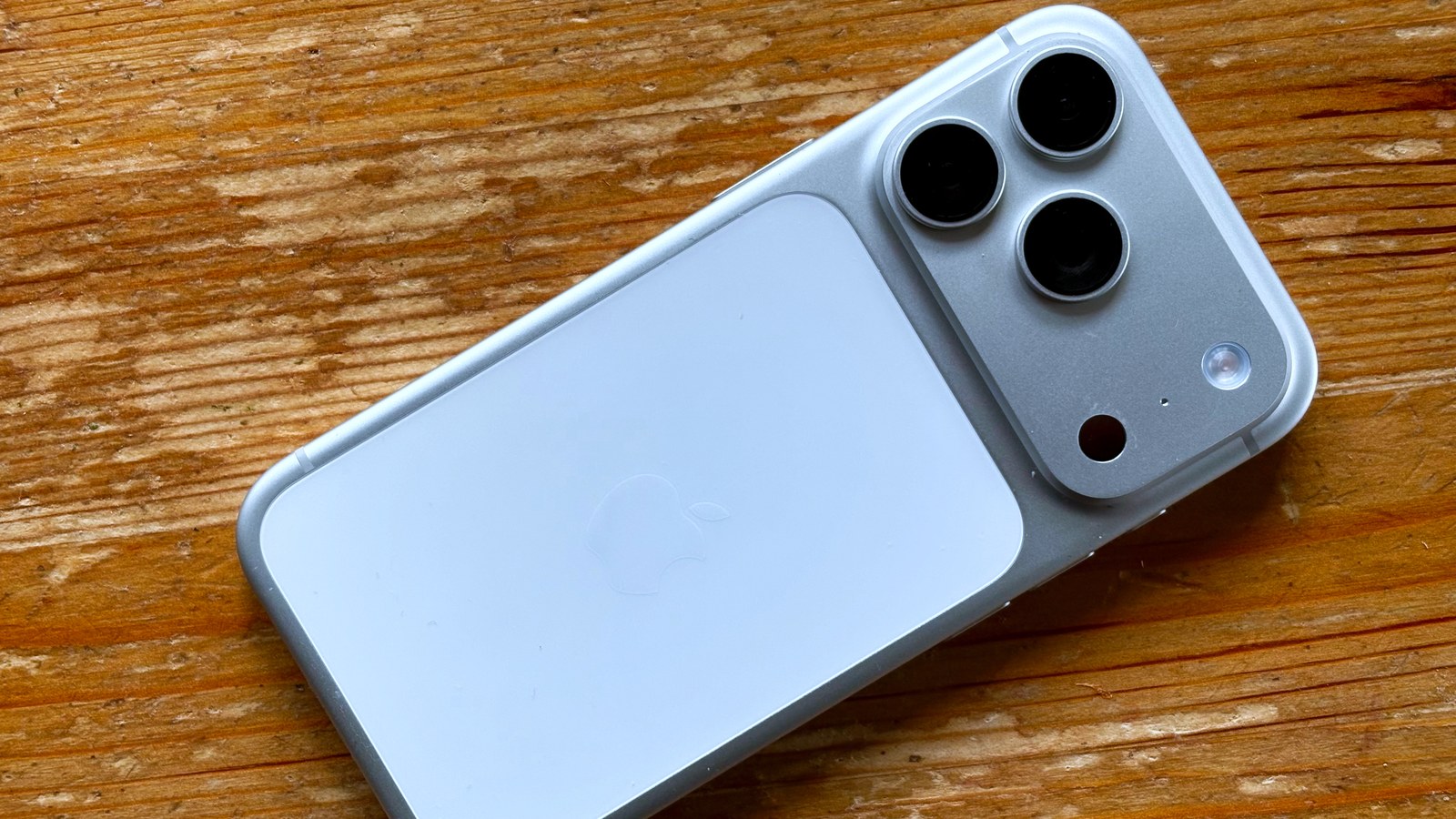 iPhone 17 Pro de cor clara, com a parte de trás voltada para cima, sobre uma mesa de madeira; vê-se o módulo com três câmaras e a maçã da Apple