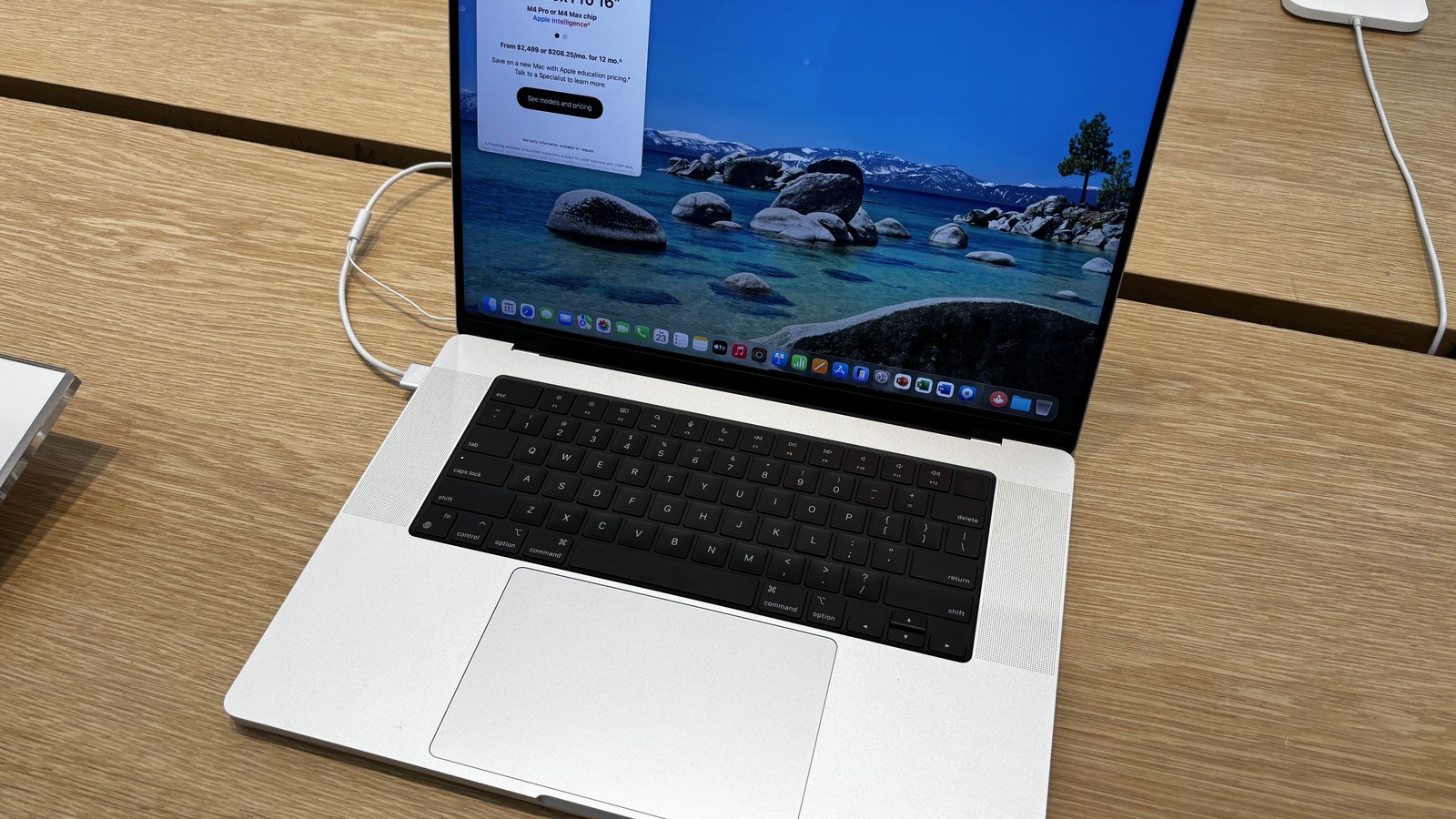 MacBook Pro de 16 polegadas, prateado, aberto sobre uma mesa de madeira, com o ecrã ligado a mostrar o ambiente de trabalho do macOS e uma janela com informação sobre o modelo