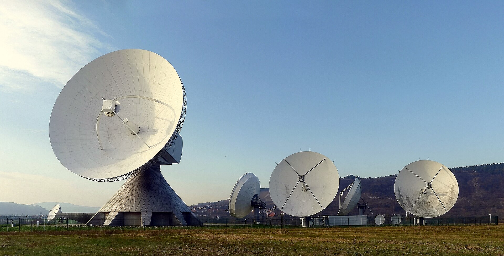 Várias antenas parabólicas brancas de grande diâmetro num campo verde, uma delas maior sobre uma base cónica, com colinas ao fundo