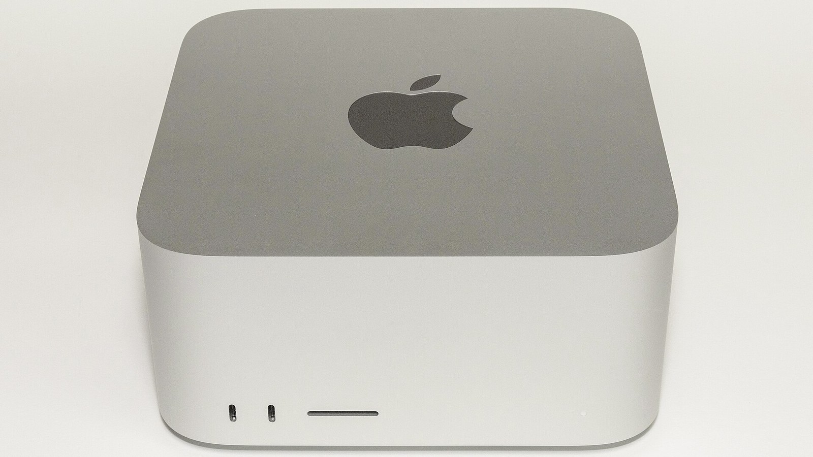 Mac Studio prateado visto de frente sobre fundo claro: corpo cúbico de cantos arredondados, maçã da Apple na face superior e, na frente, duas portas USB-C e uma ranhura para cartões SD.