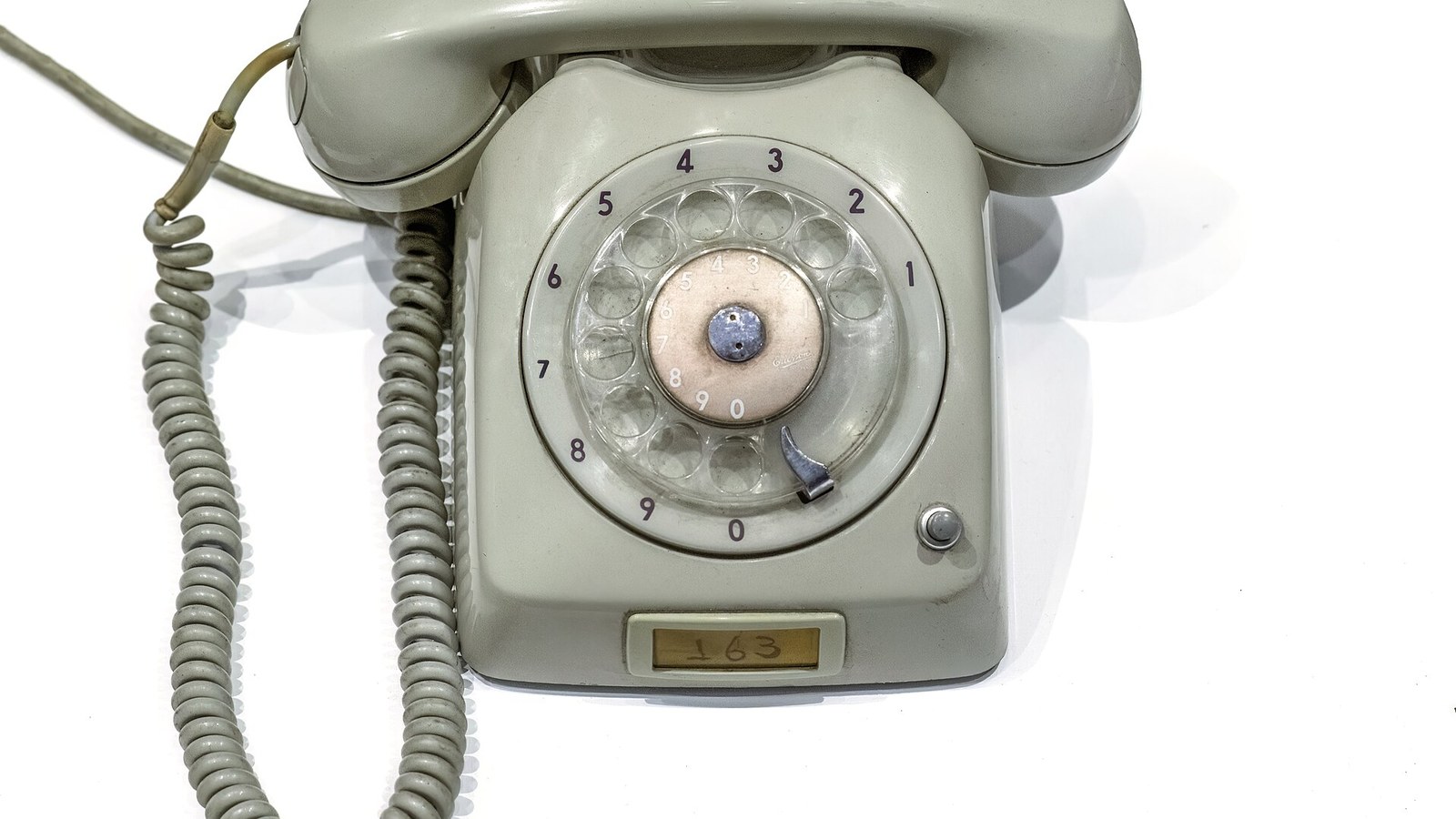 Telefone fixo de disco, cinzento, com o auscultador assente sobre a base e o fio em espiral, fotografado isolado sobre fundo branco