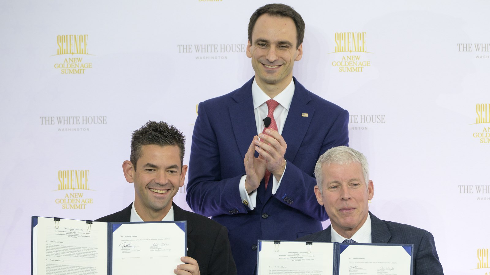Jared Isaacman, da NASA (sentado à esquerda), e Chris Wright, secretário da Energia (sentado à direita), seguram o memorando assinado; Michael Kratsios, da Casa Branca, aplaude de pé ao centro, sob um fundo com o logótipo da cimeira «Science: A New Golden Age Summit»