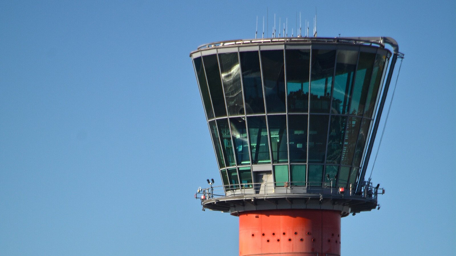 Torre de controlo do aeroporto de Heathrow, em Londres, com a cabine envidraçada e o anel inferior laranja, contra um céu azul sem nuvens