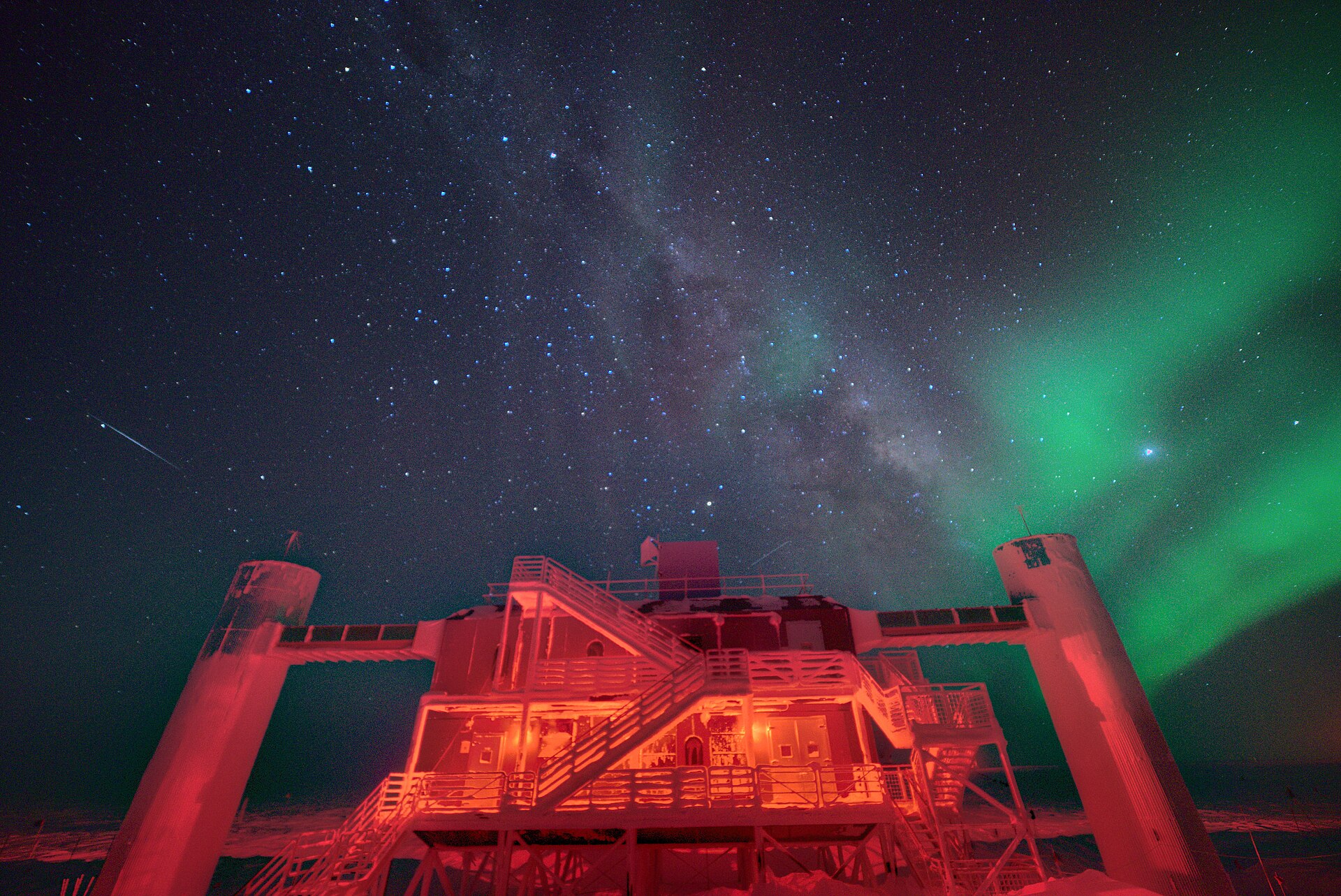 O edifício do IceCube Lab, no Pólo Sul, visto de baixo à noite, iluminado a vermelho, sob um céu estrelado com a Via Láctea ao centro e uma aurora austral verde à direita