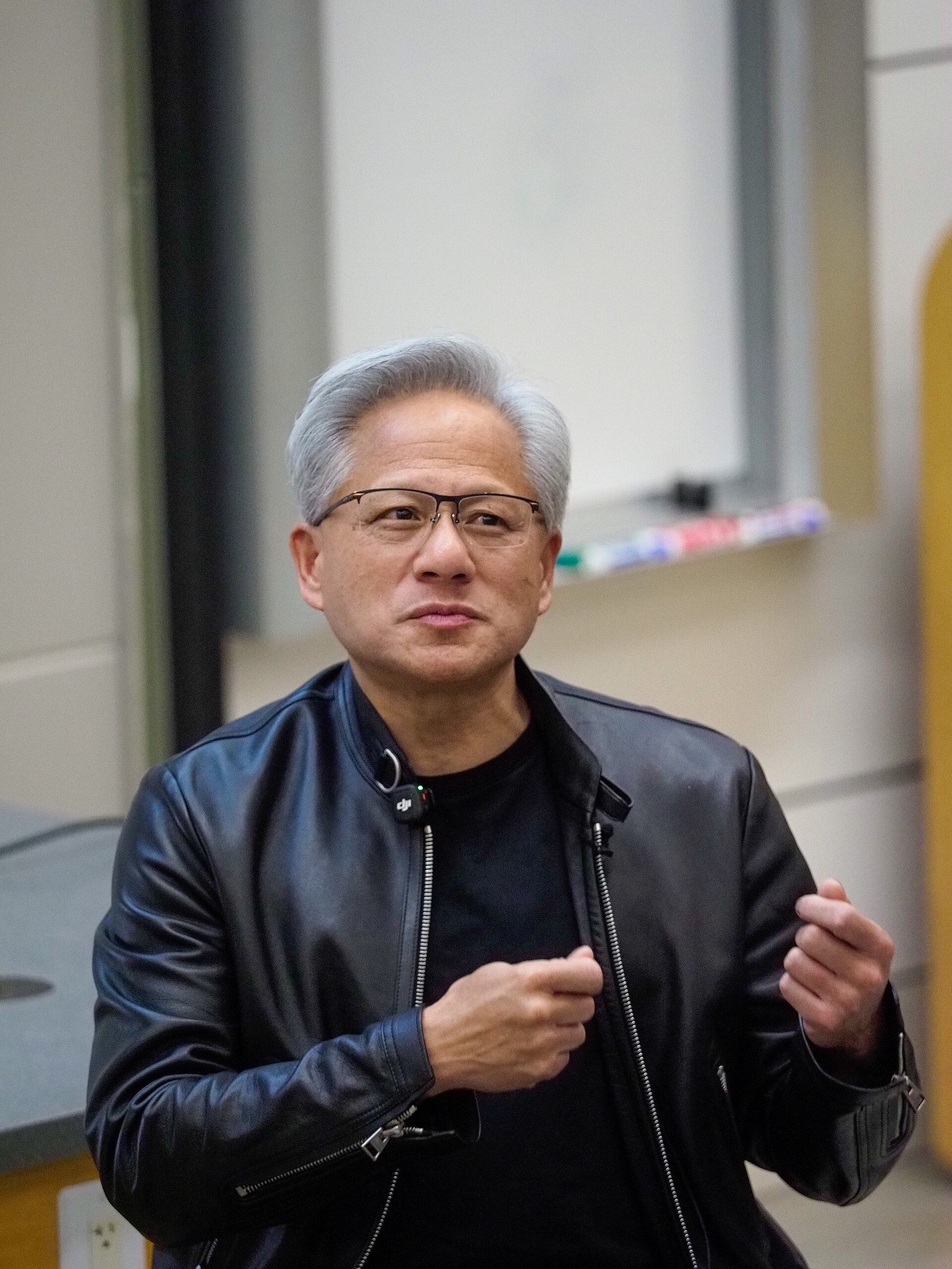 Jensen Huang, de casaco de cabedal preto e óculos, fala de pé com as mãos à altura do peito, diante de um quadro branco