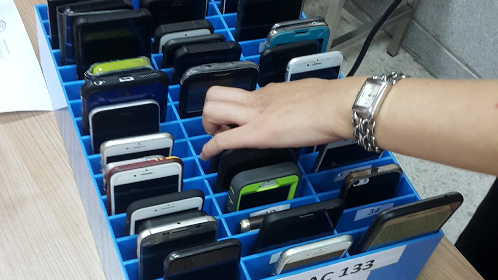 Compartimento azul com dezenas de telemóveis guardados em casas numeradas, sobre uma mesa; uma mão pousa num dos aparelhos