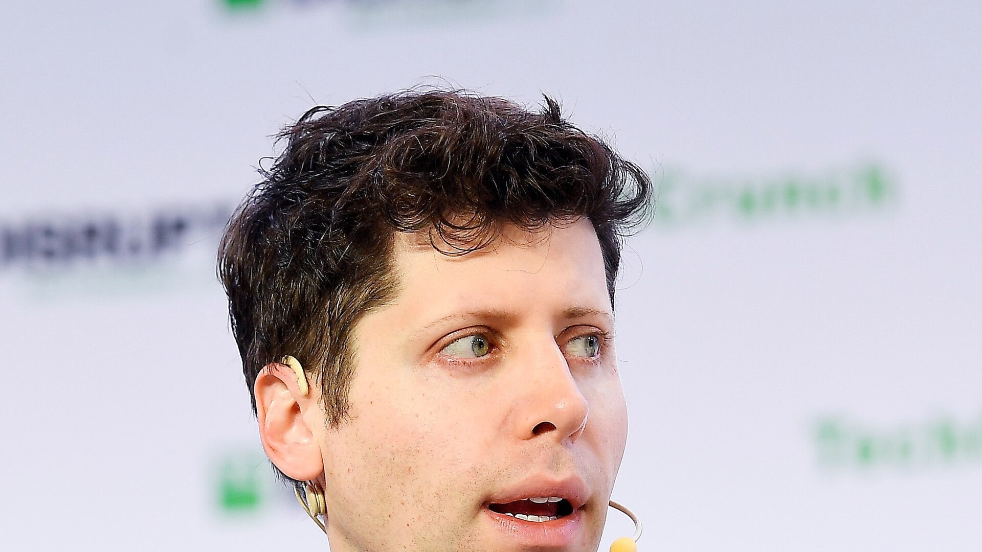 Primeiro plano de Sam Altman, cabeça e início dos ombros, cabelo ondulado escuro e camisa em malha azul, com microfone de cabeça amarelo, a olhar para a direita num palco de conferência; fundo claro desfocado com patrocínios