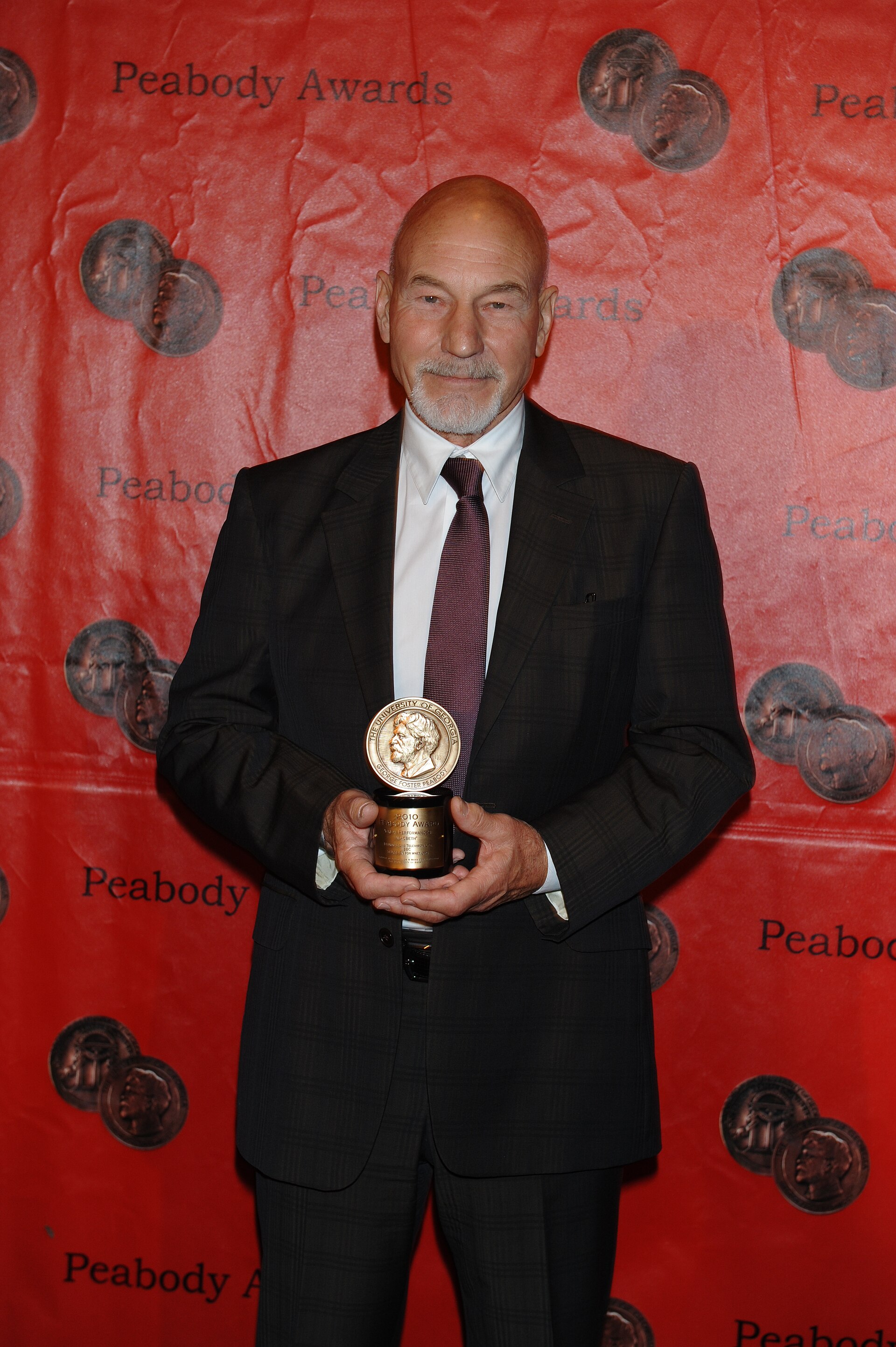 Patrick Stewart de fato escuro e gravata, segurando um prémio Peabody diante de um fundo vermelho