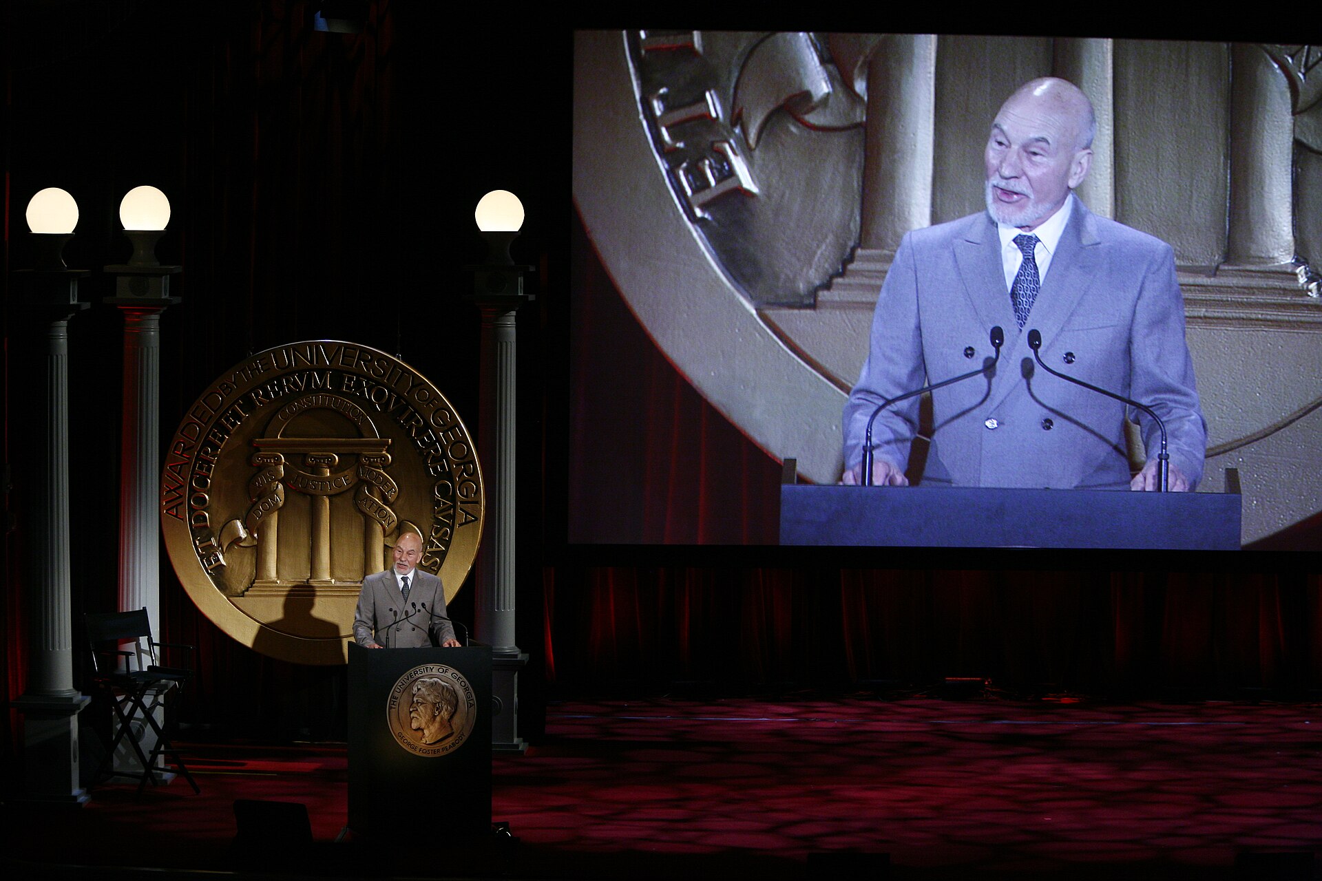 Patrick Stewart fala num púlpito numa cerimónia dos Peabody Awards, com a sua imagem projectada num grande ecrã