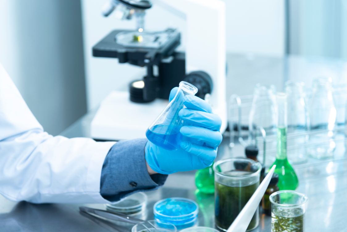 Investigador em laboratório com jaleco e luvas a analisar amostra líquida azul num erlenmeyer, com microscópio ao fundo