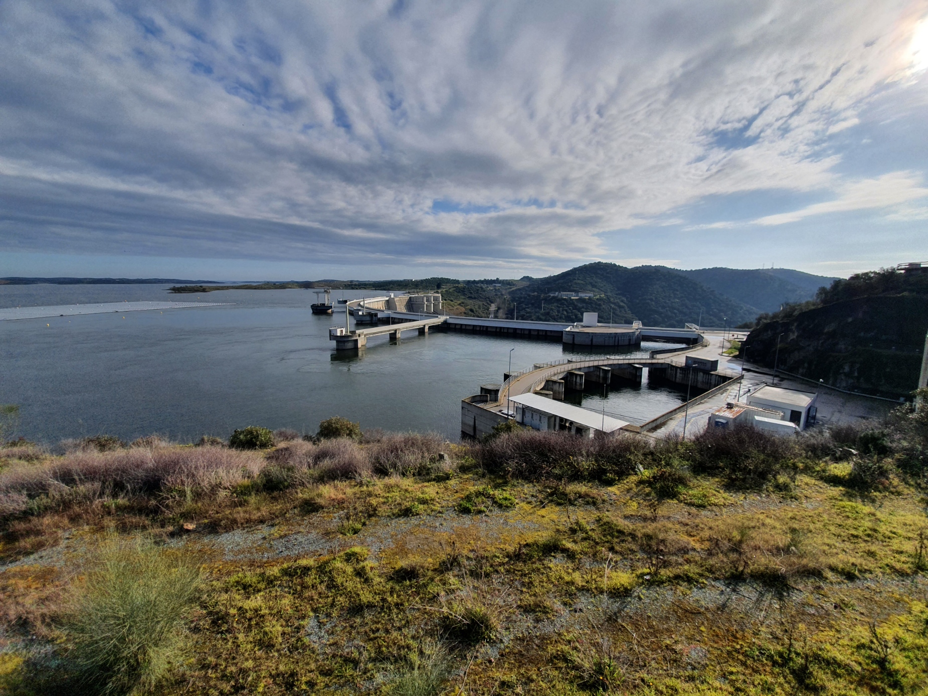 Vista sobre a barragem de Alqueva: plano de água do reservatório em primeiro plano, com a barragem, o descarregador e as estruturas da central no lado direito, encostas cobertas de vegetação rasteira à volta e céu muito nublado