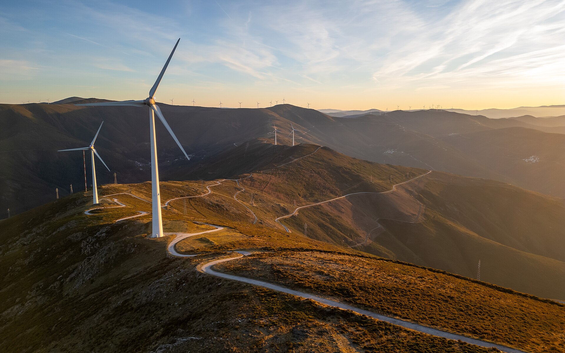 Aerogeradores numa crista de montanha ao pôr do sol, com várias torres eólicas distribuídas pelas encostas e caminhos de acesso em terra batida