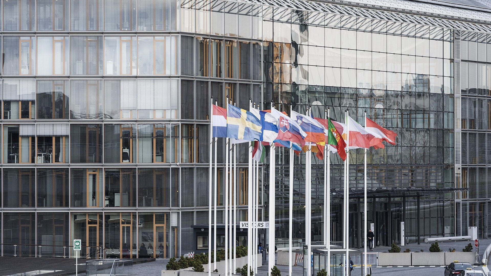 Fachada de vidro da sede do Grupo BEI no Kirchberg, em Luxemburgo, com bandeiras de Estados-membros da União Europeia alinhadas à entrada