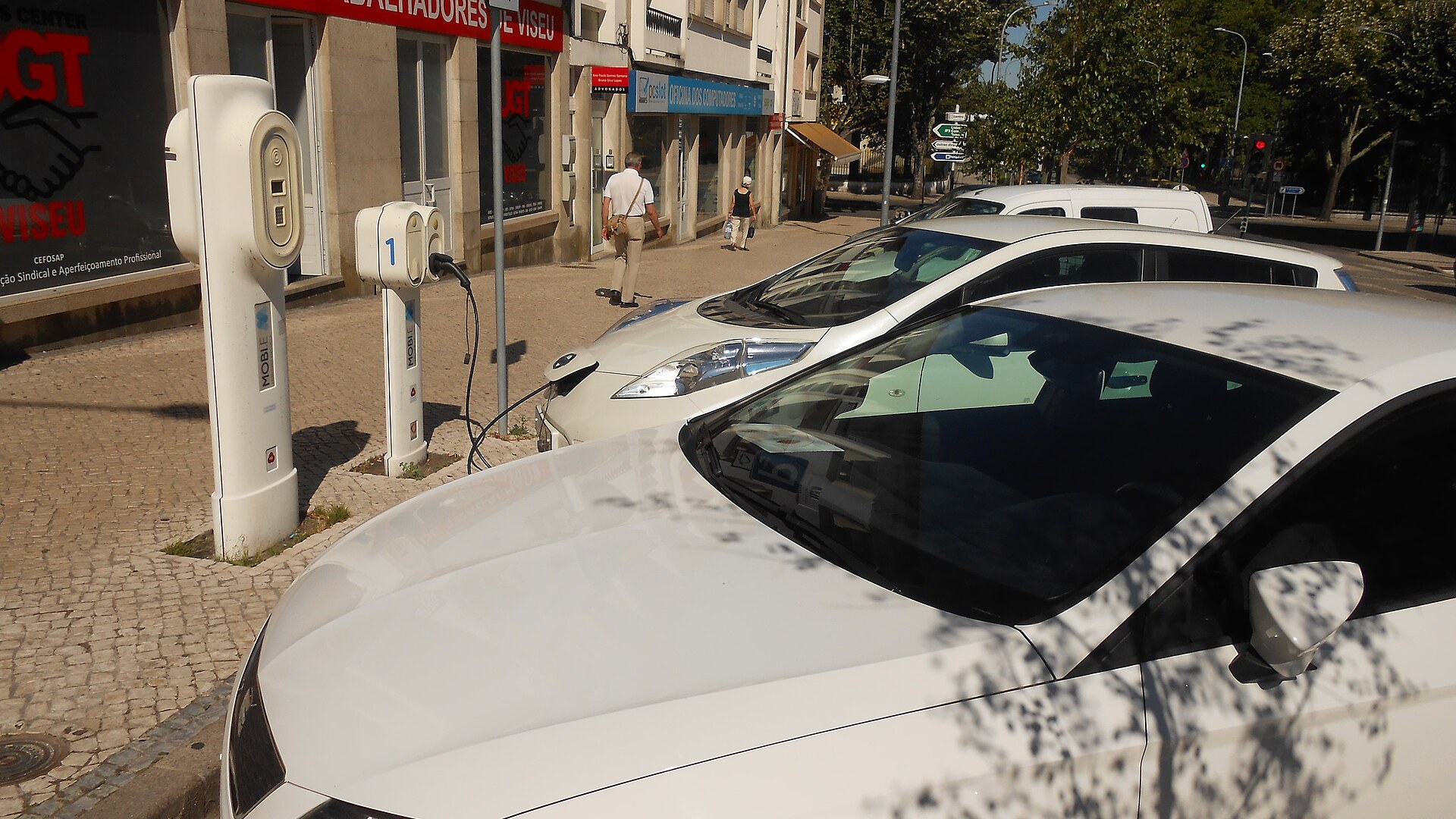 Dois automóveis elétricos brancos estacionados junto a postos de carregamento MOBI.E numa rua de Viseu, com uma pessoa a passar no passeio e edifícios comerciais ao fundo