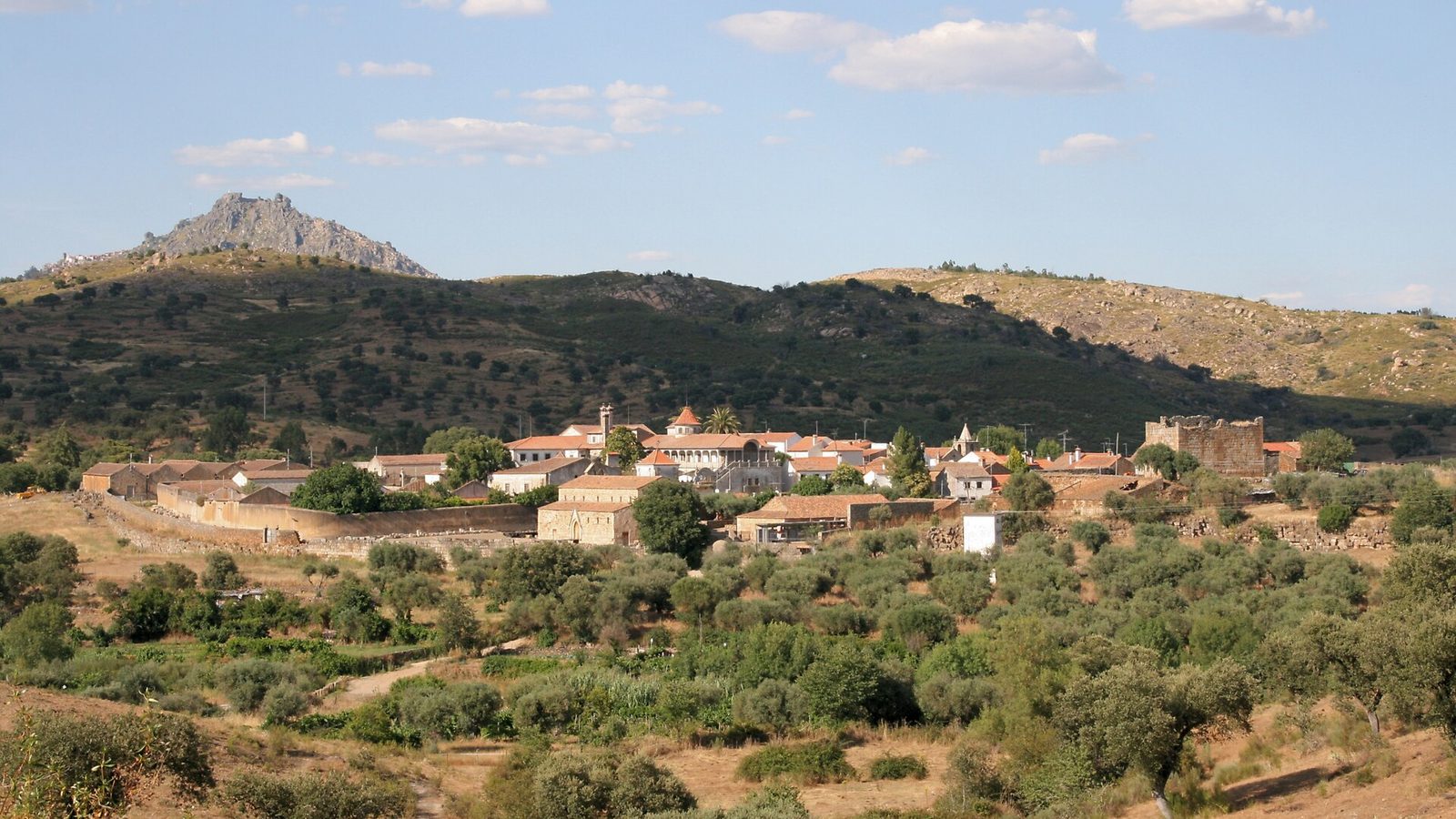 Paisagem de encostas graníticas com olival em primeiro plano, uma aldeia de casas brancas com telhado de telha no meio e, ao fundo à esquerda, um cabeço rochoso com um povoado no topo, sob céu azul com nuvens.