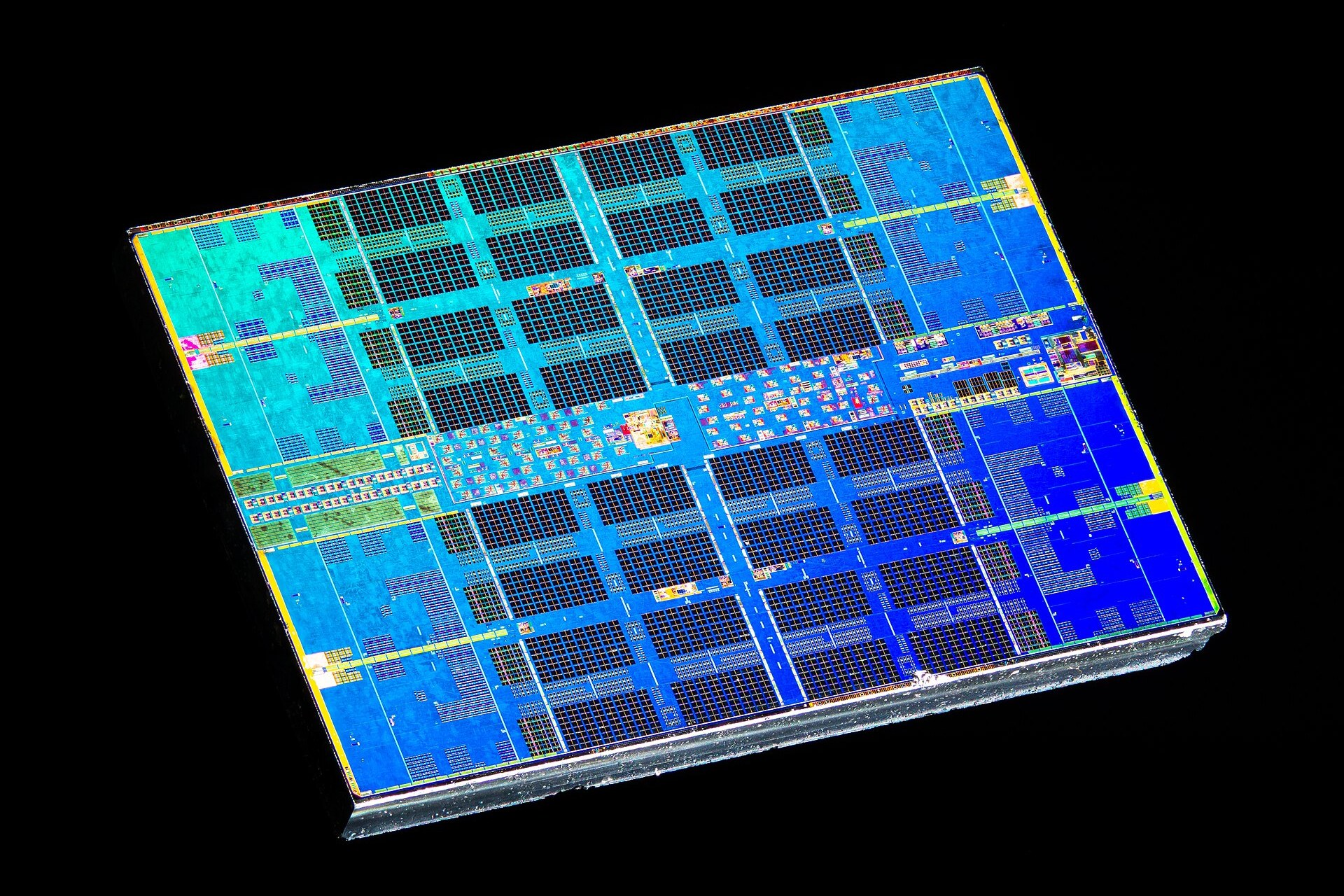 Fotografia microscópica da pastilha de um processador: uma área rectangular inclinada sobre fundo preto, preenchida por blocos de circuitos em azul e turquesa dispostos numa grelha regular