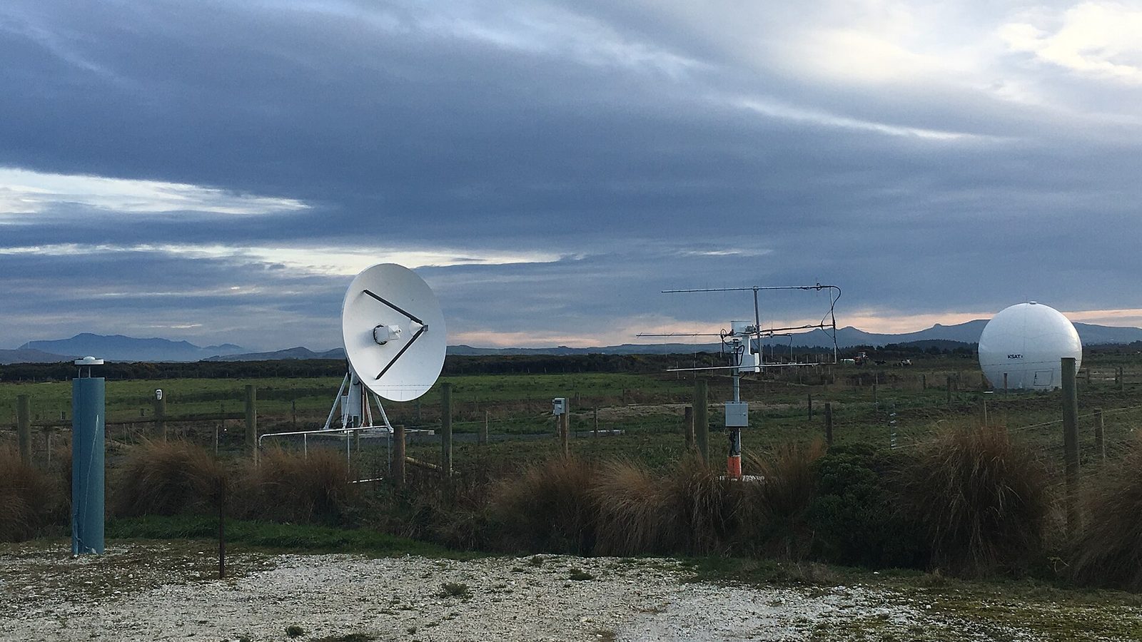 Estação terrestre de satélites num campo aberto: uma antena parabólica branca apontada ao céu, uma antena de dipolos sobre um mastro e um radomo branco redondo ao fundo, sob um céu carregado de nuvens escuras.