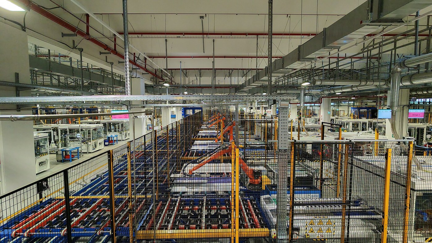 Interior de uma fábrica com uma linha de produção longa e automatizada: tapetes com recipientes azuis e vermelhos, braços robóticos laranja dentro de grades de protecção amarelas e, ao fundo, um corredor com painéis e ecrãs de controlo.