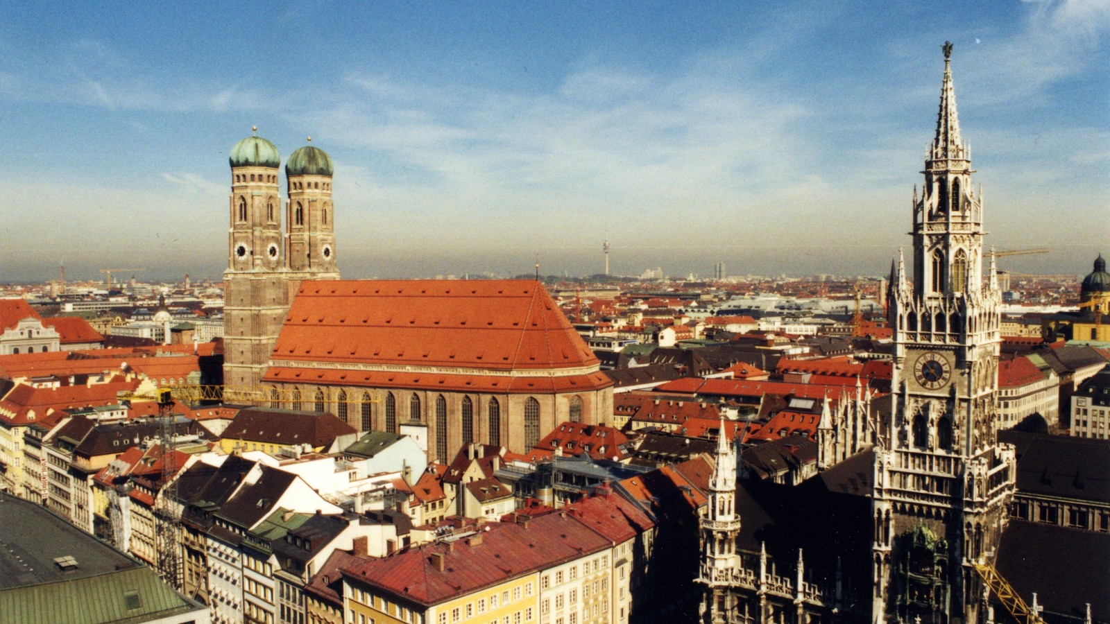 Vista sobre o centro de Munique: à esquerda a catedral Frauenkirche, com duas torres de cúpulas verdes, e à direita a torre do novo paço municipal, sobre telhados vermelhos.